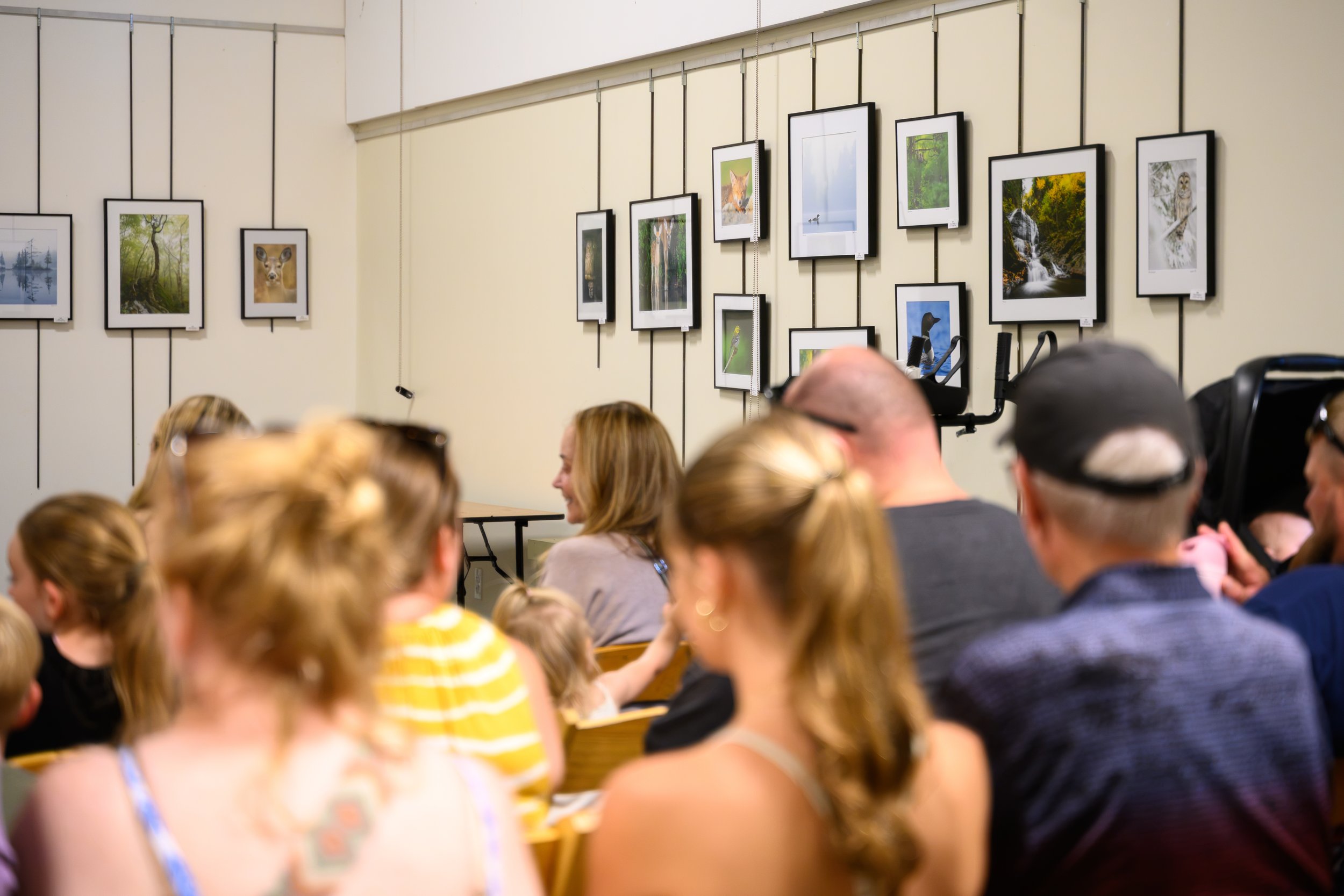 An indoor art gallery with hanging framed nature photographs on a beige wall, and a group of people seated, possibly attending a presentation or event.