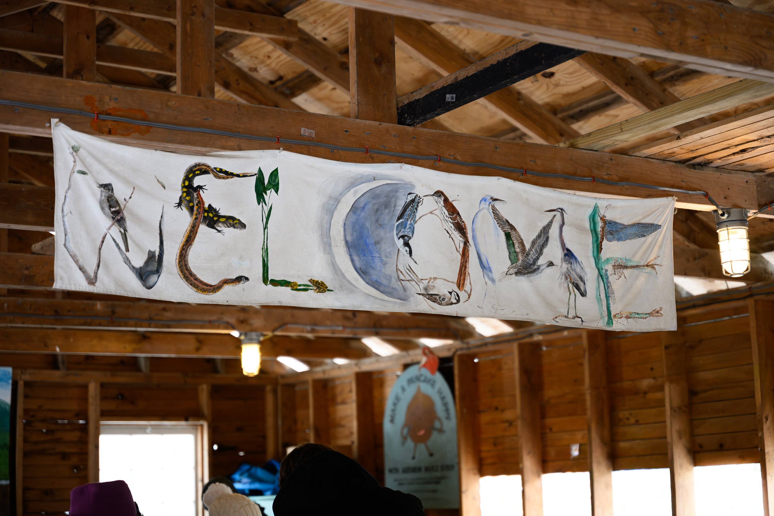 A banner with painted illustrations of various birds and a snake, hanging from a wooden ceiling structure in a rustic interior.