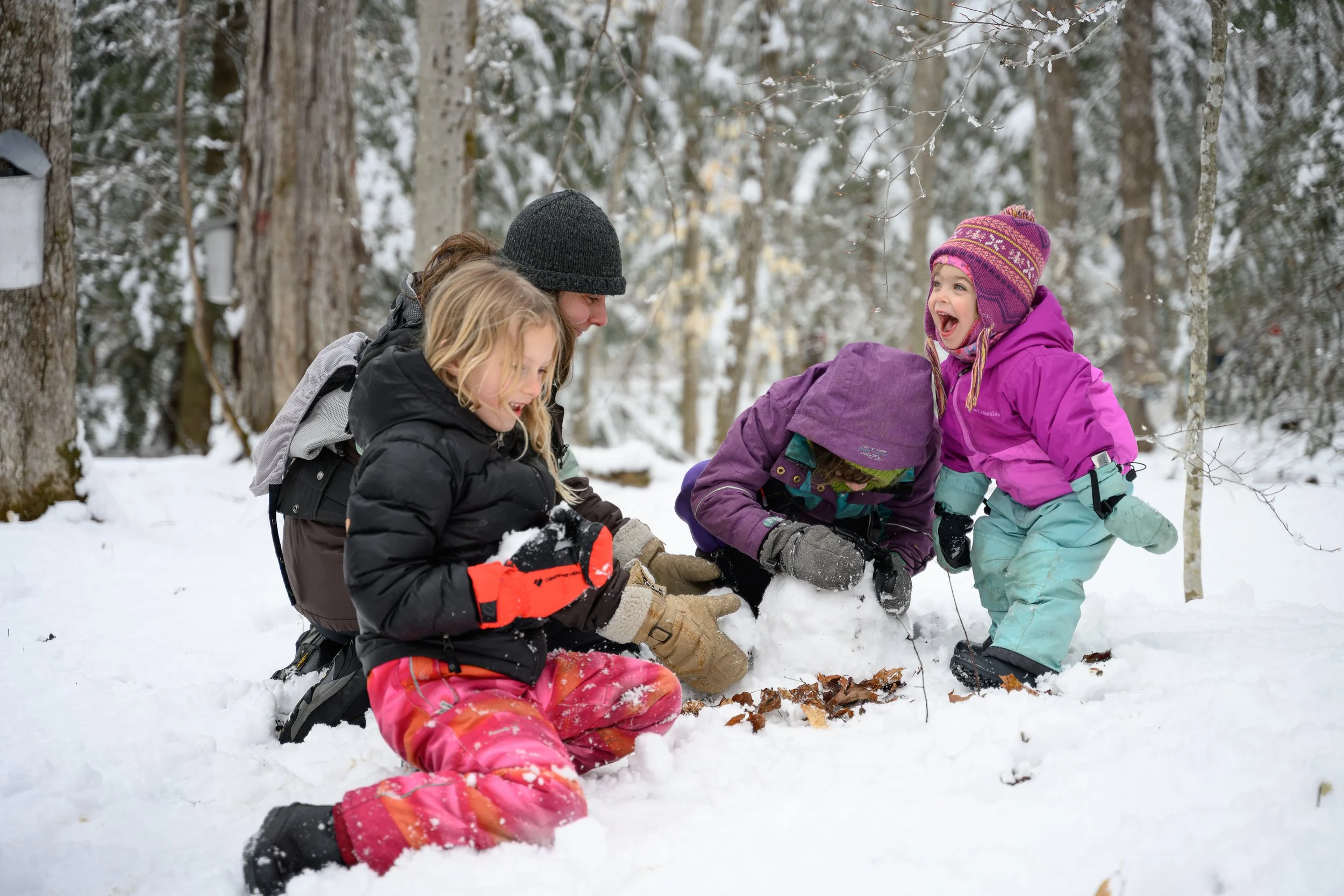 A group of children and a woman playing in the snow in a wooded area, wearing winter clothing, engaged in snowy outdoor activity.