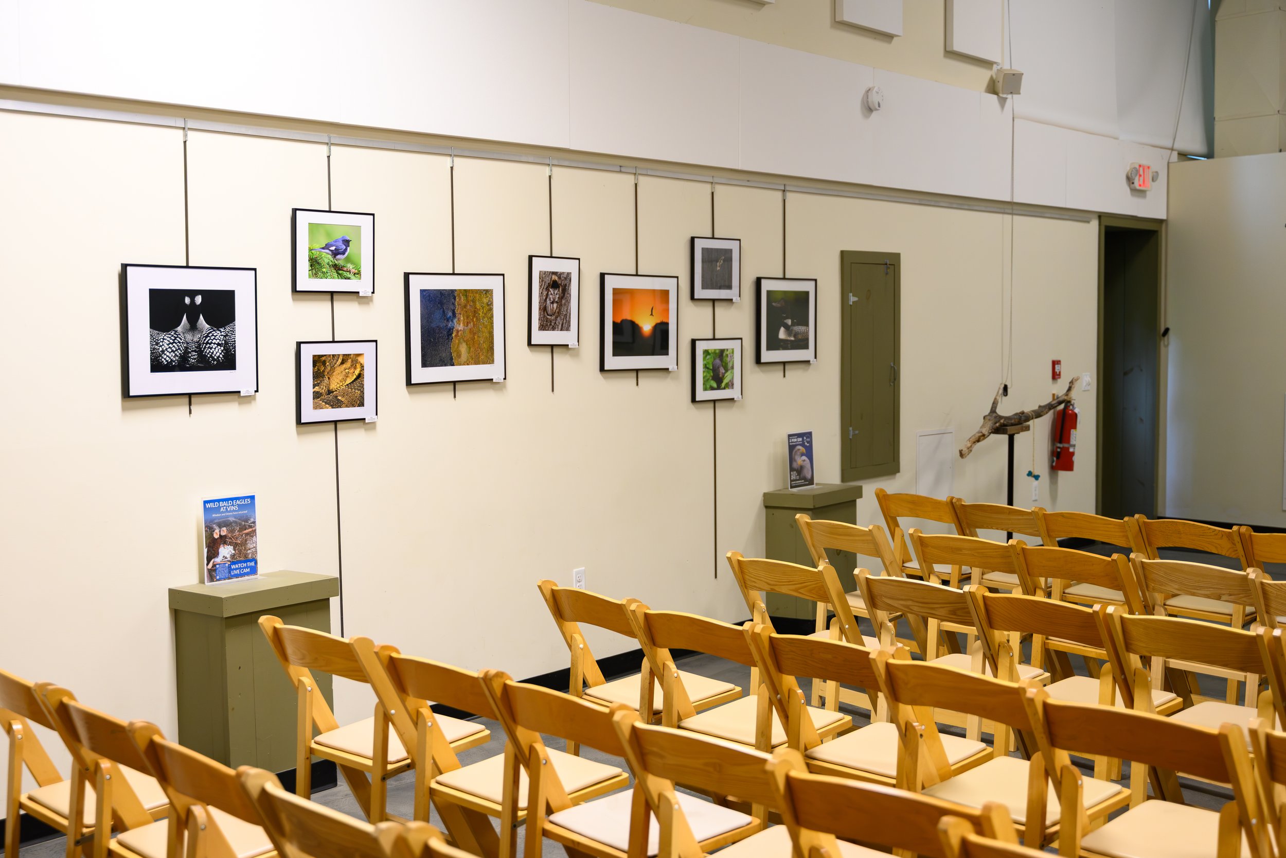 An art gallery with framed photographs on the wall, rows of wooden chairs facing the gallery, and informational signs on the tables.