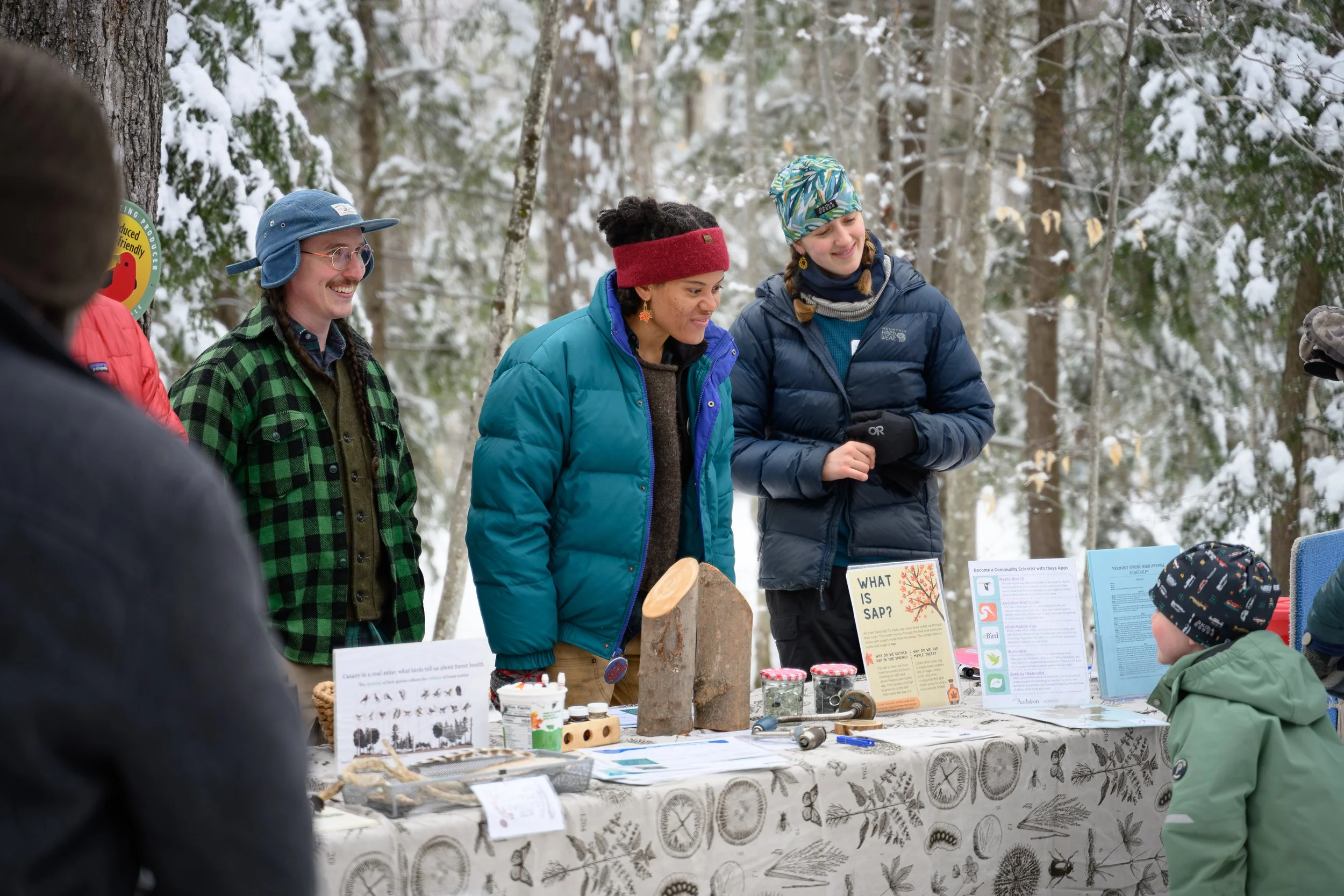A group of four women in winter clothing gathered behind a table outdoors in a snowy forest, engaging in conversation and smiling. The table displays informational materials, signs, and various small items.
