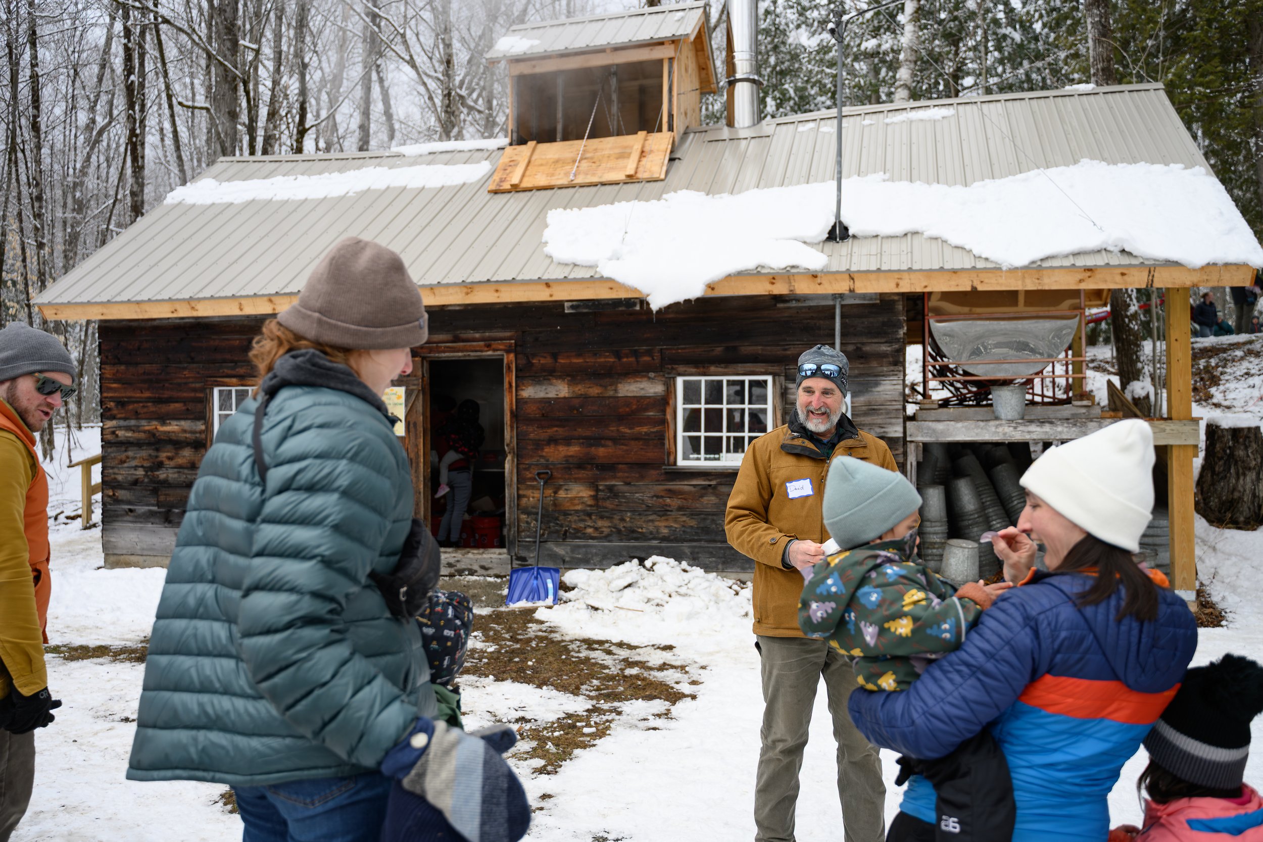 Group of people gathered outdoors in front of a rustic wooden cabin with snow on the ground and on the cabin roof, engaging in conversation on a winter day.