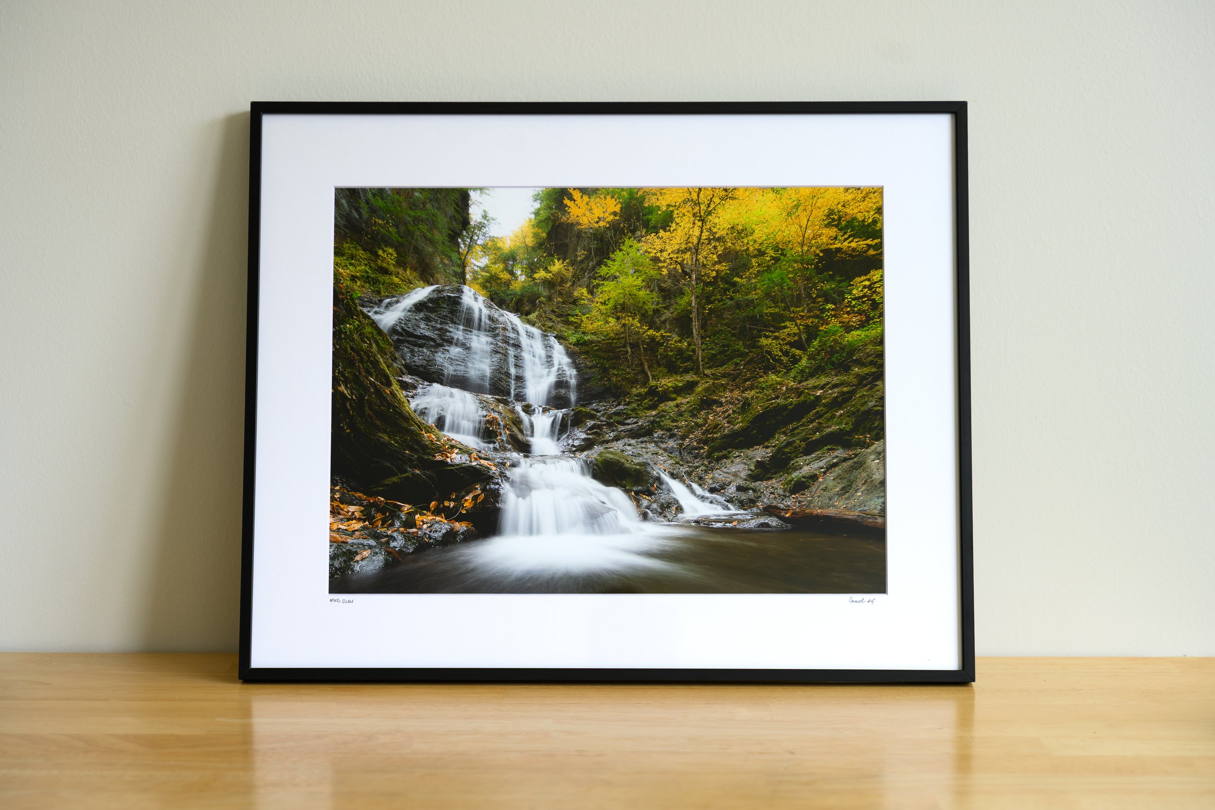 Framed landscape photograph of a waterfall flowing through a forest with yellow and green foliage, displayed on a wall.
