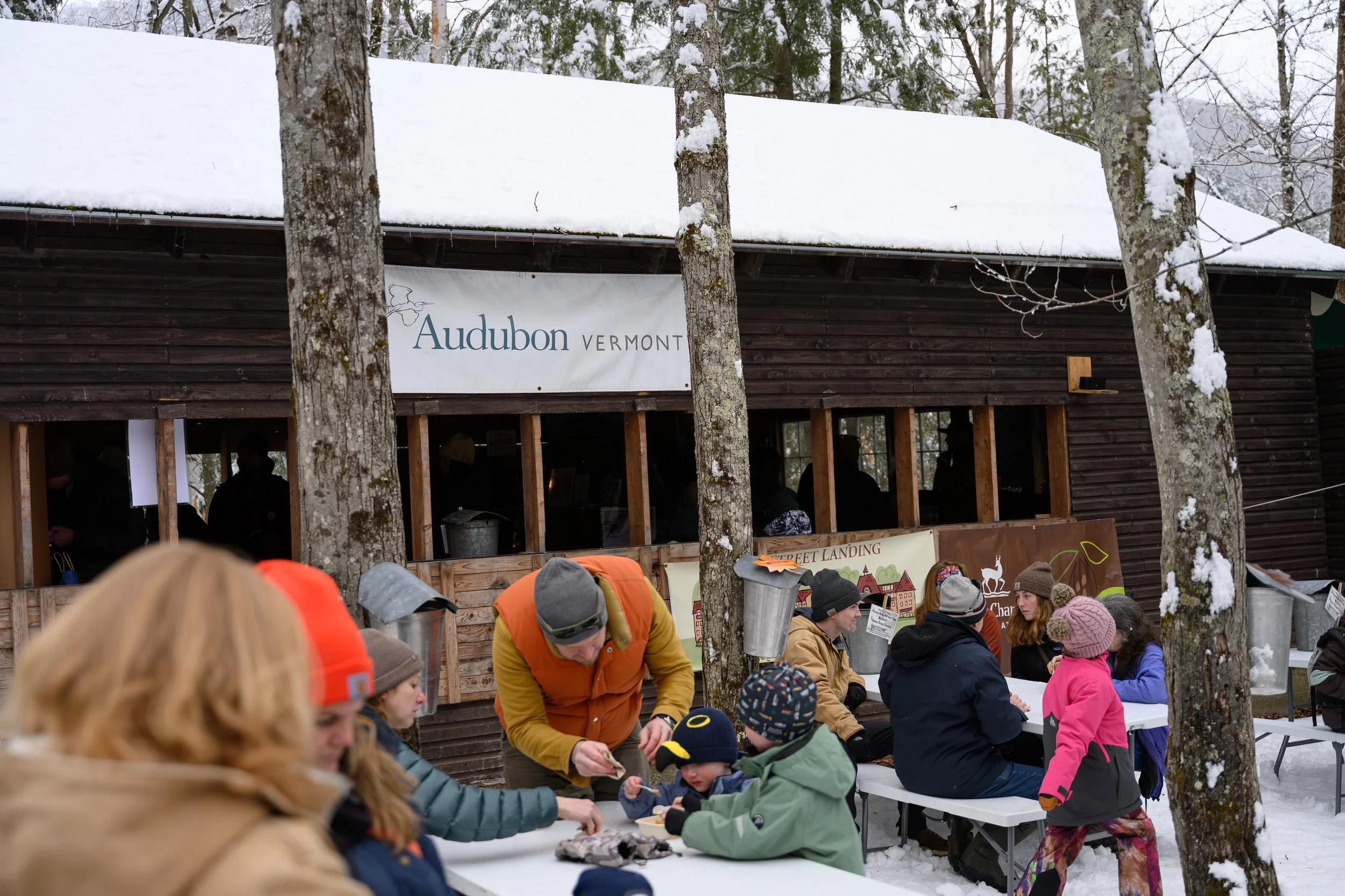 People gather outdoors in winter near a wooden building with a sign that reads "Audubon Vermont". Snow covers the roof, and trees with snow-covered branches are in the background. Some people are seated at tables, while others are standing and intera