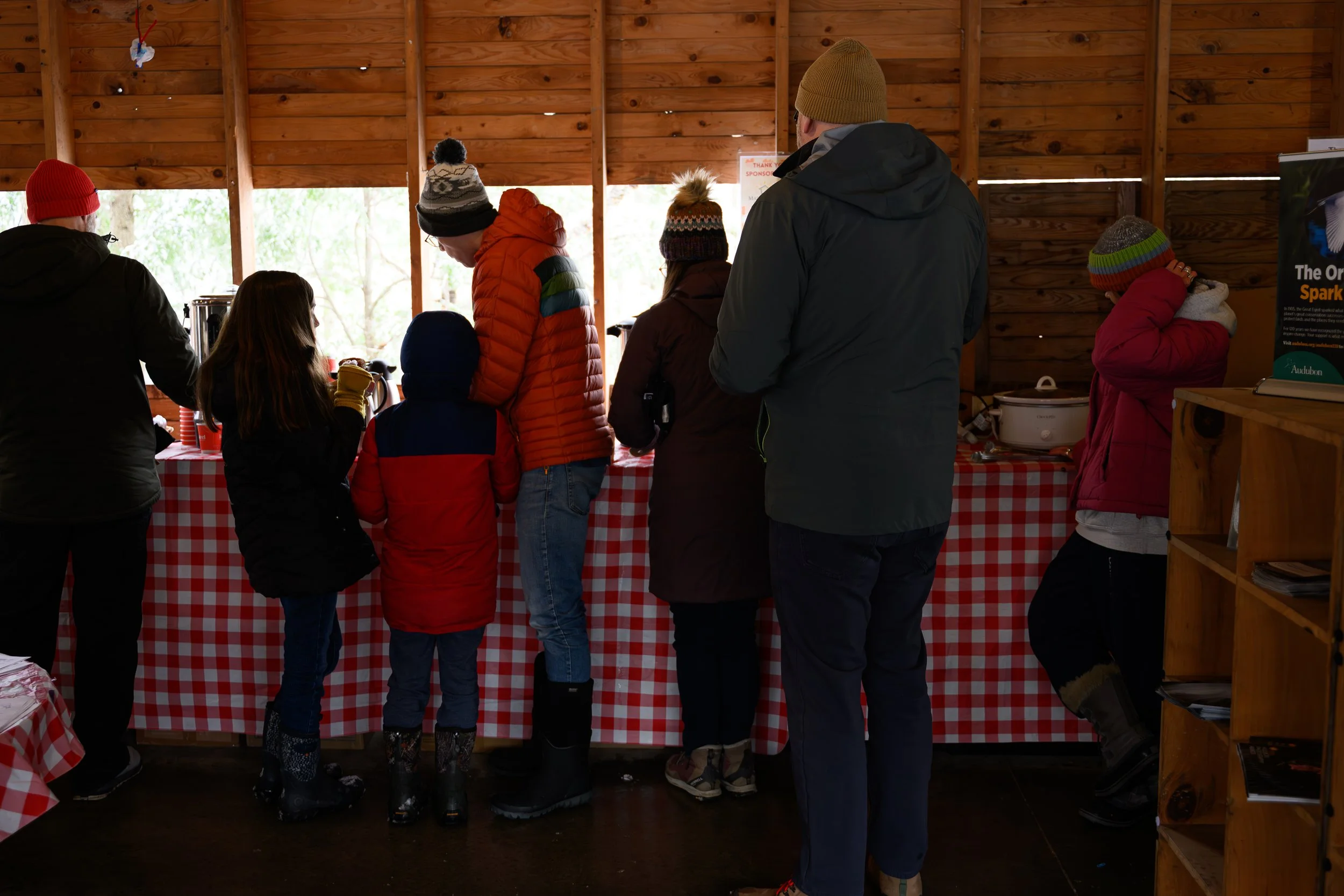 People gathered inside a wooden shelter with a red checkered tablecloth, participating in a food or drink tasting event, dressed in winter clothing.
