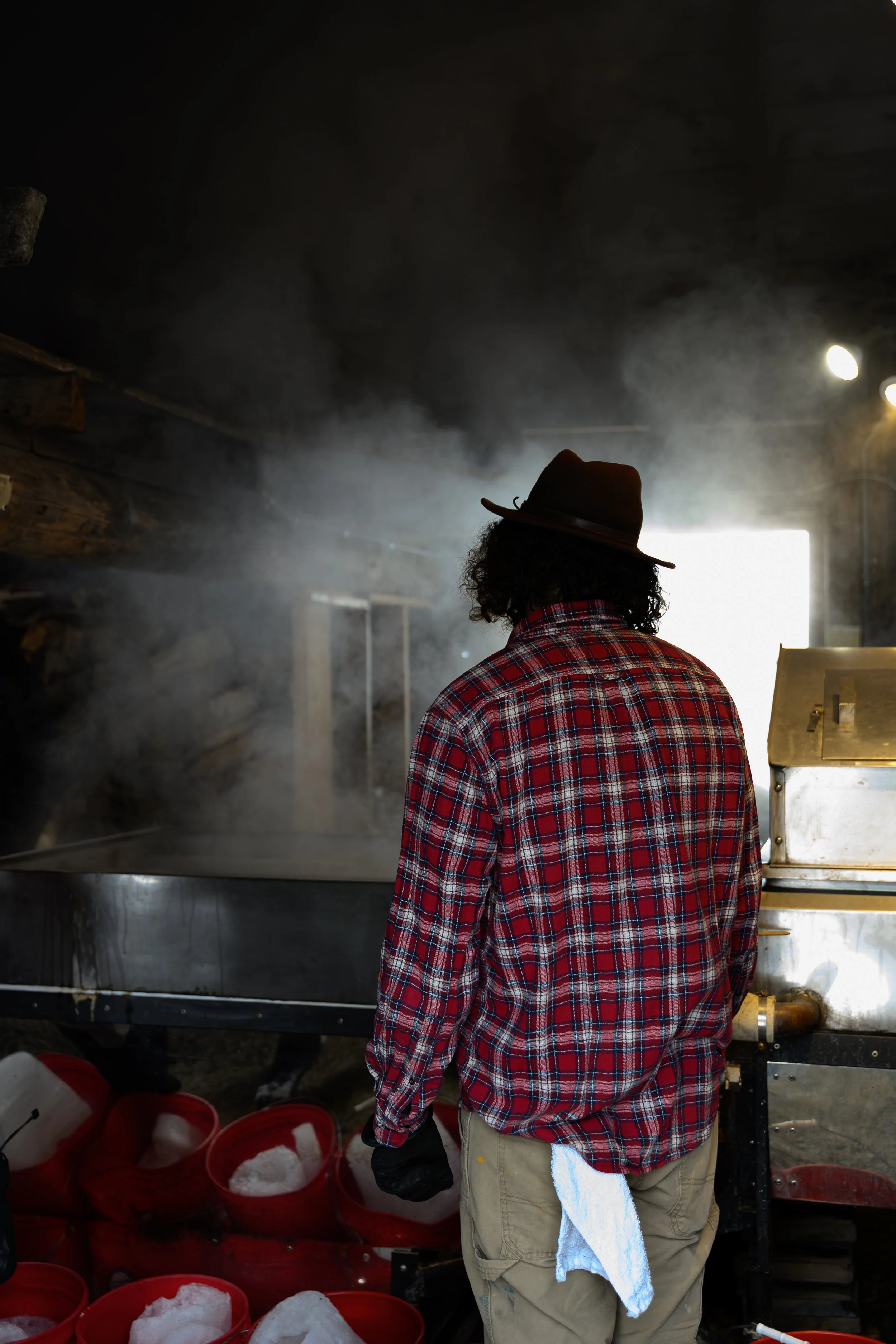 A person with curly hair wearing a brown hat, red plaid shirt, and beige pants stands in front of a stove with steam rising, in a rustic kitchen. Several red buckets filled with ice are on the floor.