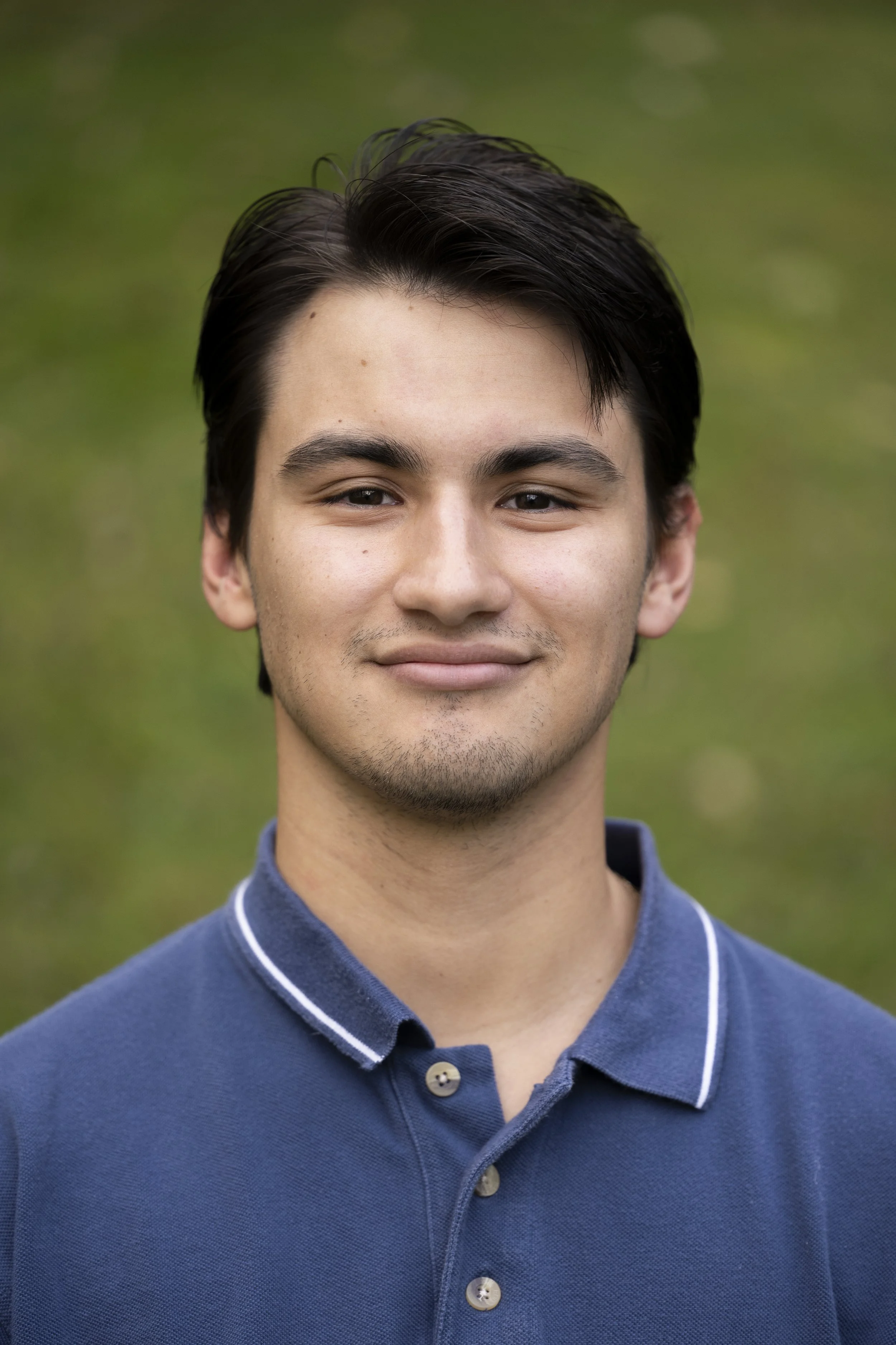 A young man with dark hair wearing a blue collared shirt smiling at the camera in an outdoor setting.