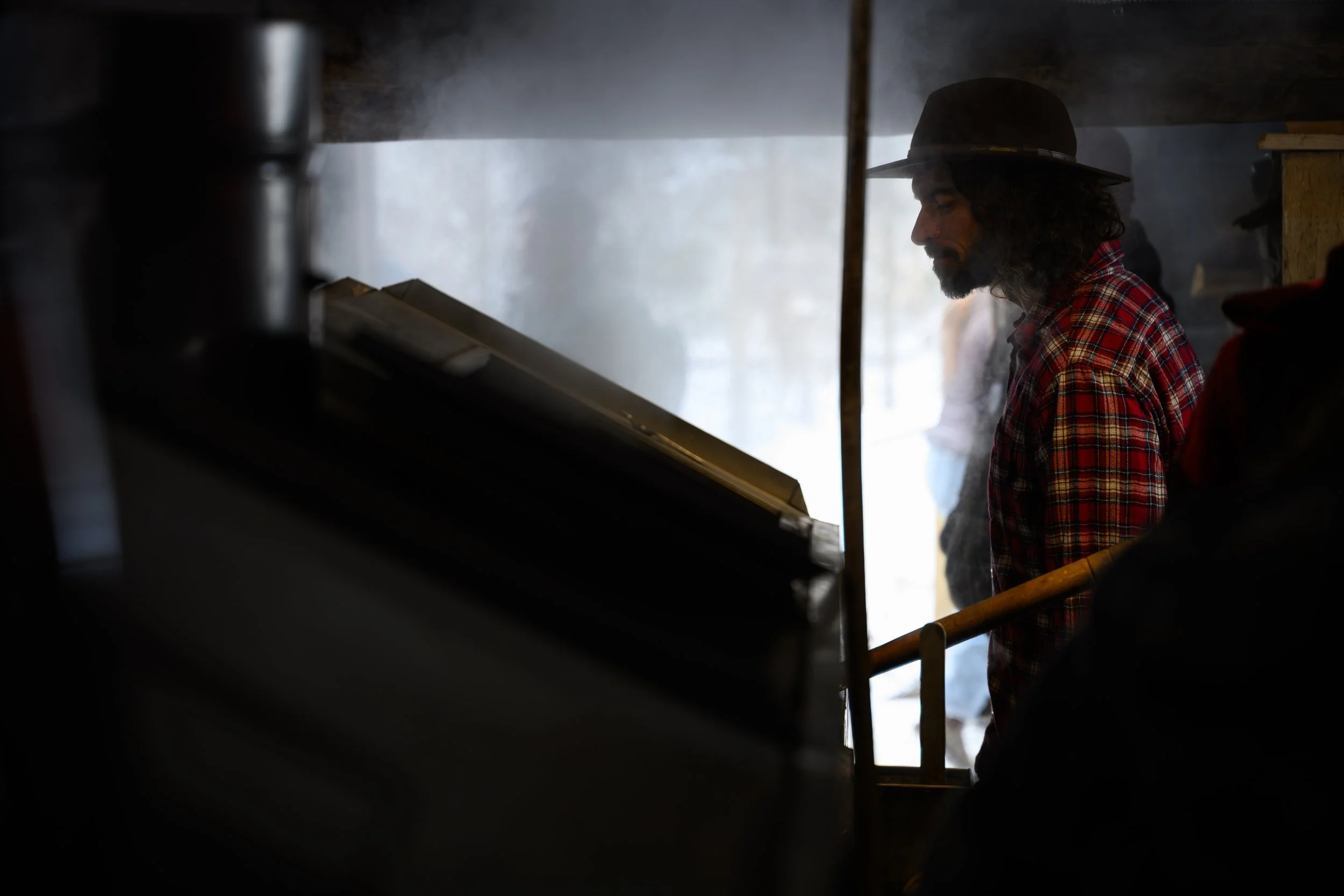 A man with long curly hair and a beard wearing a black hat and red plaid shirt standing in a smoky, dimly lit room.