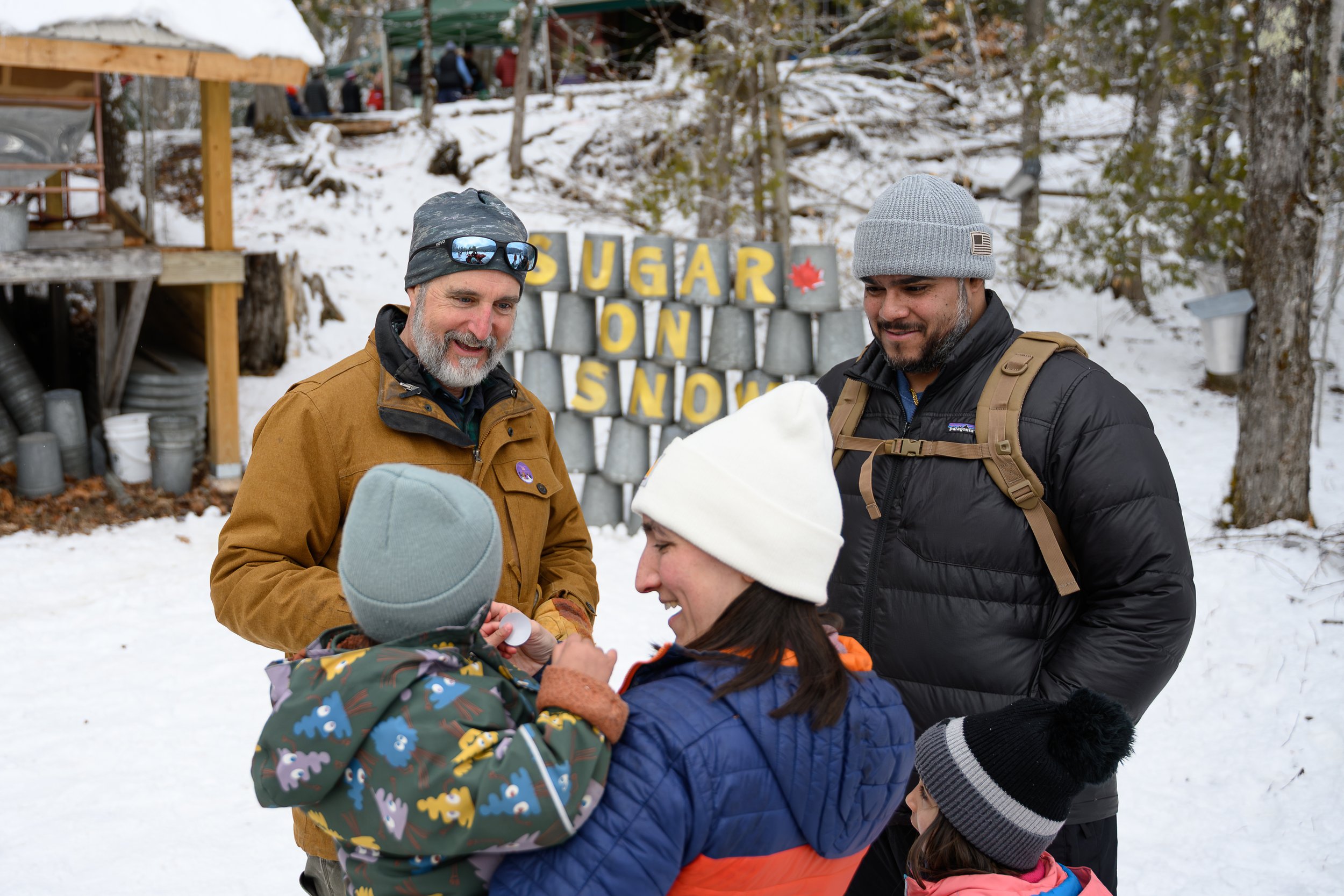 Two men and two children outdoors in snowy weather, with a sign reading 'SUGAR ON SNOW' in the background. The men are dressed warmly, one wearing a gray knit hat and a black jacket, and the other with a gray beanie and sunglasses. The children are w