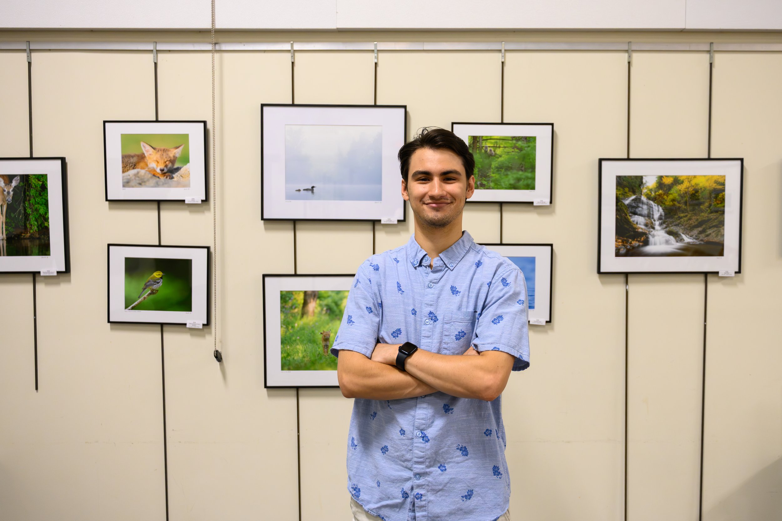 A young man with dark hair smiling, wearing a light blue shirt with a floral pattern, standing in front of framed nature photographs at an art exhibit.