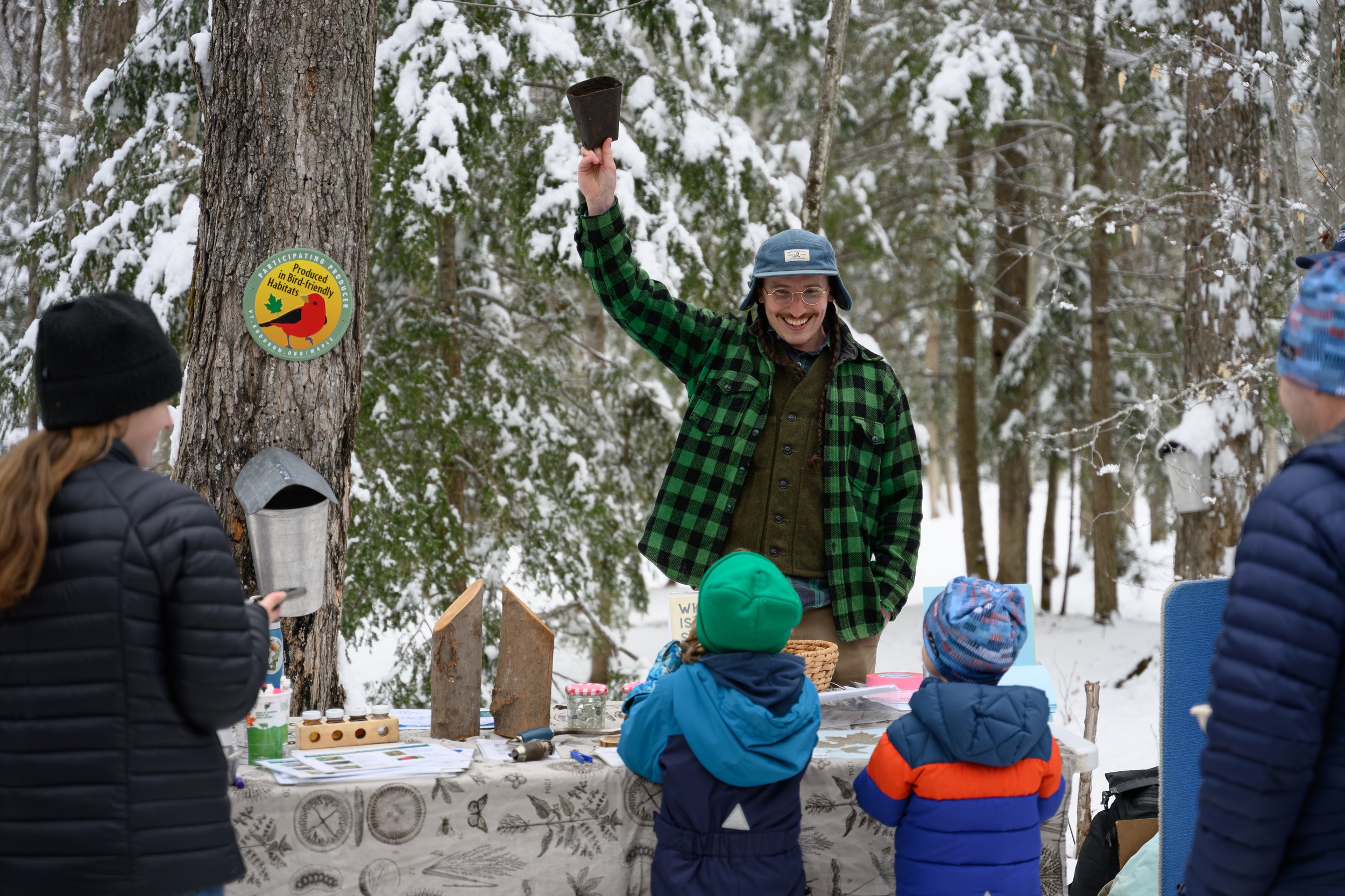 A man smiling and wearing glasses, a blue hat, and a green and black plaid jacket, raising his hand holding a black object in a snowy outdoor forest. Several children wearing winter jackets and hats are gathered around a table with informational mate