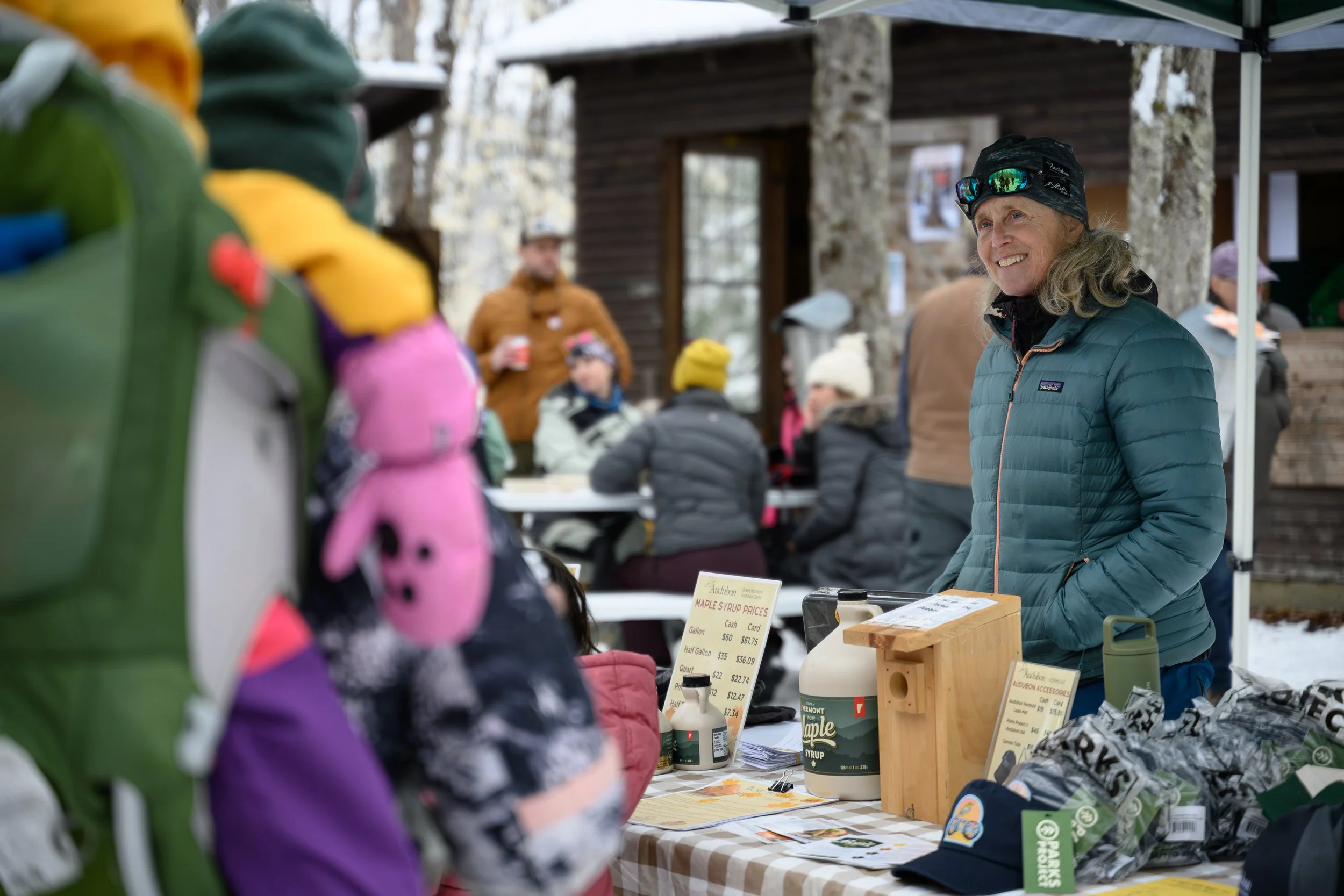 A smiling woman in a blue jacket and black cap with sunglasses on her head at an outdoor market stall, with other people in the background and items on display such as maple syrup bottles, caps, and informational signs.