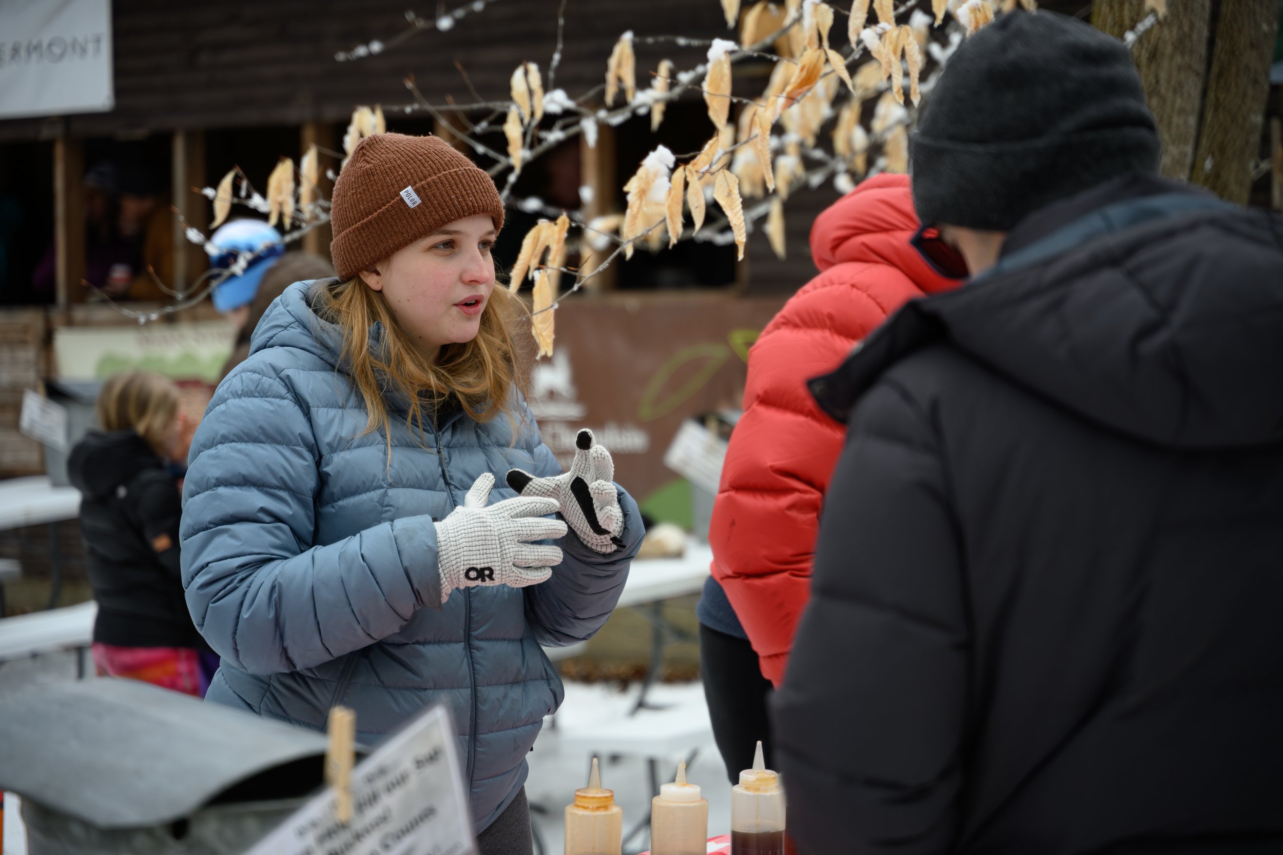 A young woman wearing a brown beanie and a light blue puffer jacket talks to a man wearing a dark beanie and red jacket at an outdoor market stall with various bottles of sauces and condiments on display. In the background, there are other people and