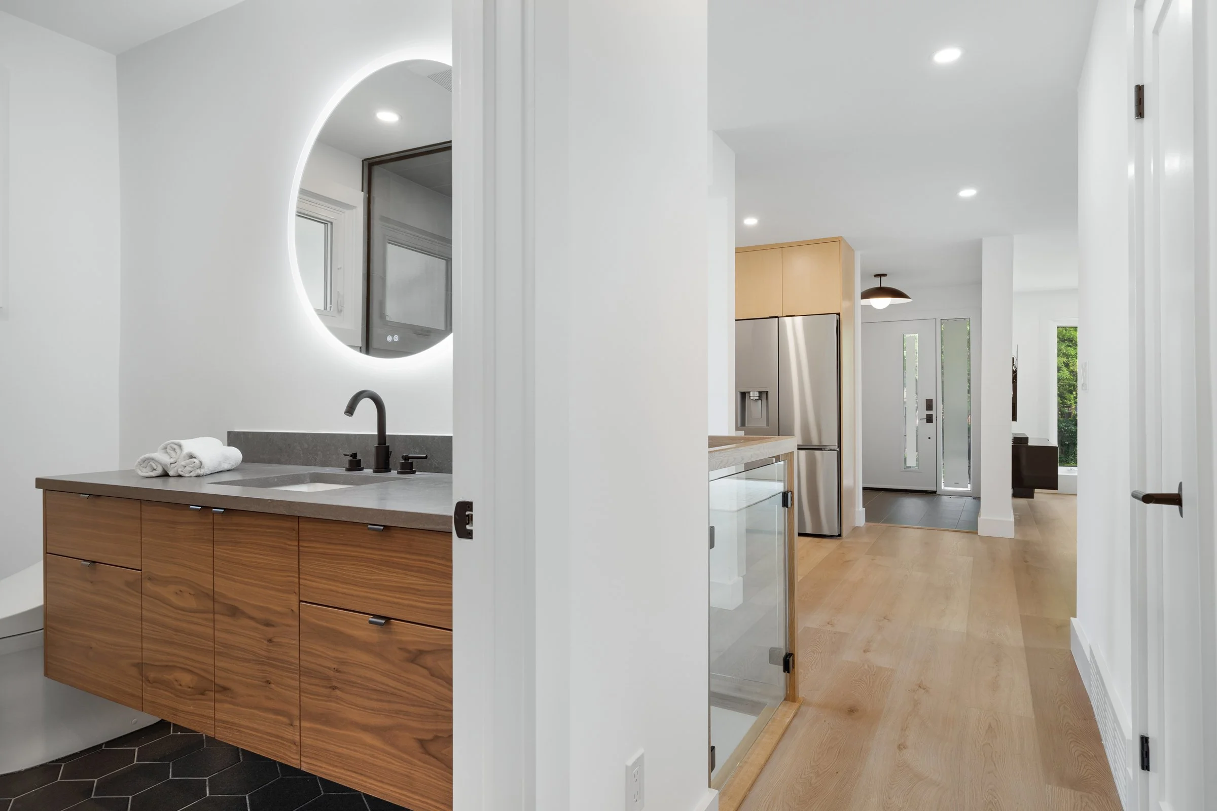 Modern interior with a bathroom and kitchen. The bathroom has a wooden vanity, round backlit mirror, and dark fixtures. The kitchen features a stainless steel refrigerator. The space is bright with wood flooring and recessed lighting.