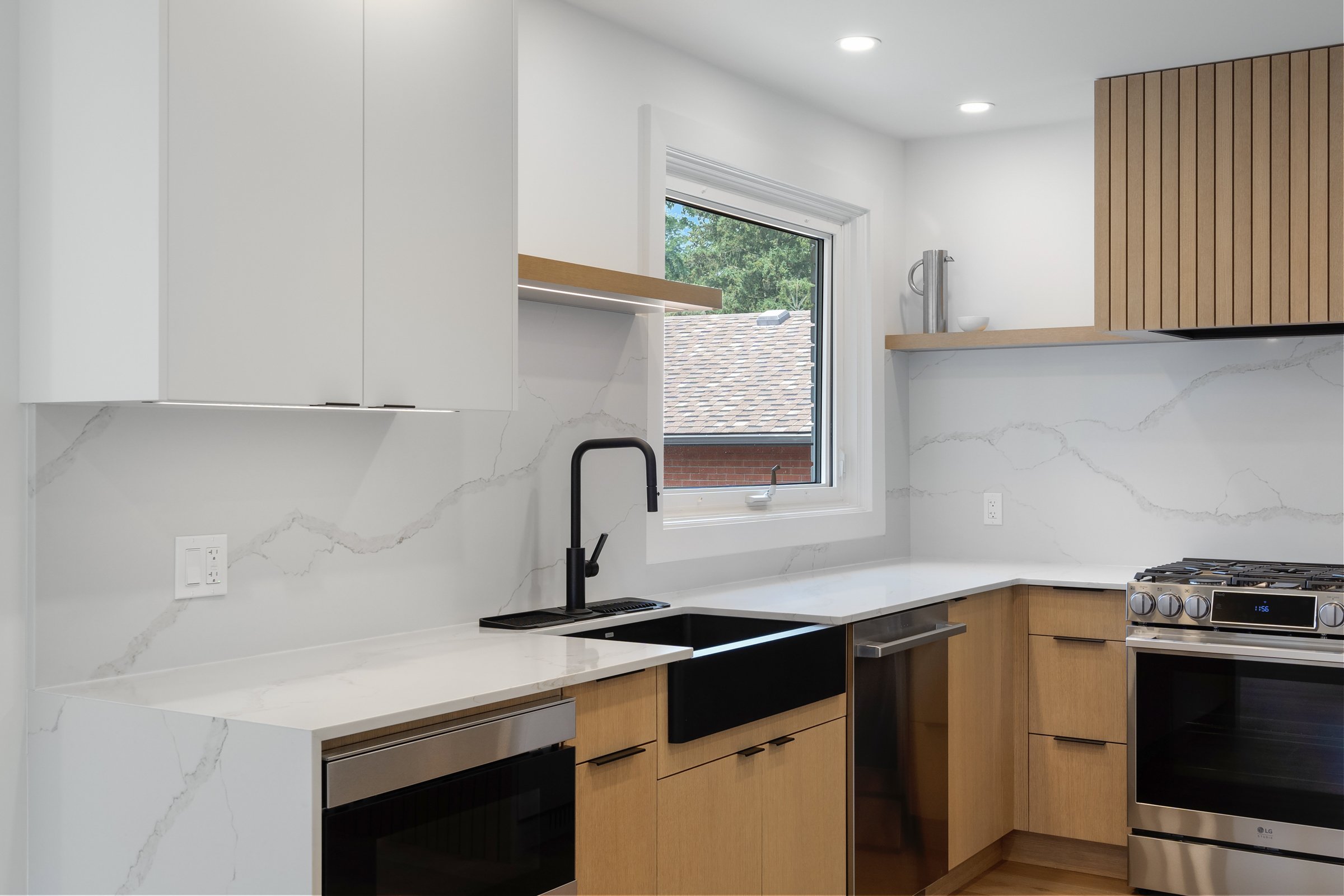 Modern kitchen with white and wooden cabinetry, black faucet, marble countertops, stainless steel appliances, and a window.