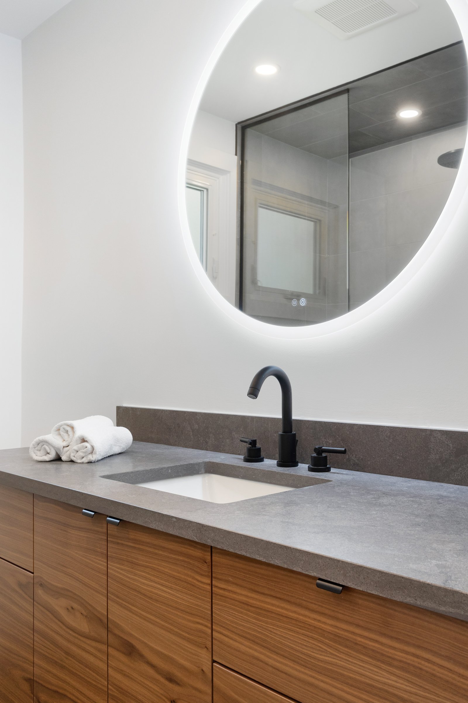 Modern bathroom vanity with round backlit mirror, wooden cabinets, gray countertop, black faucet, and rolled white towels.