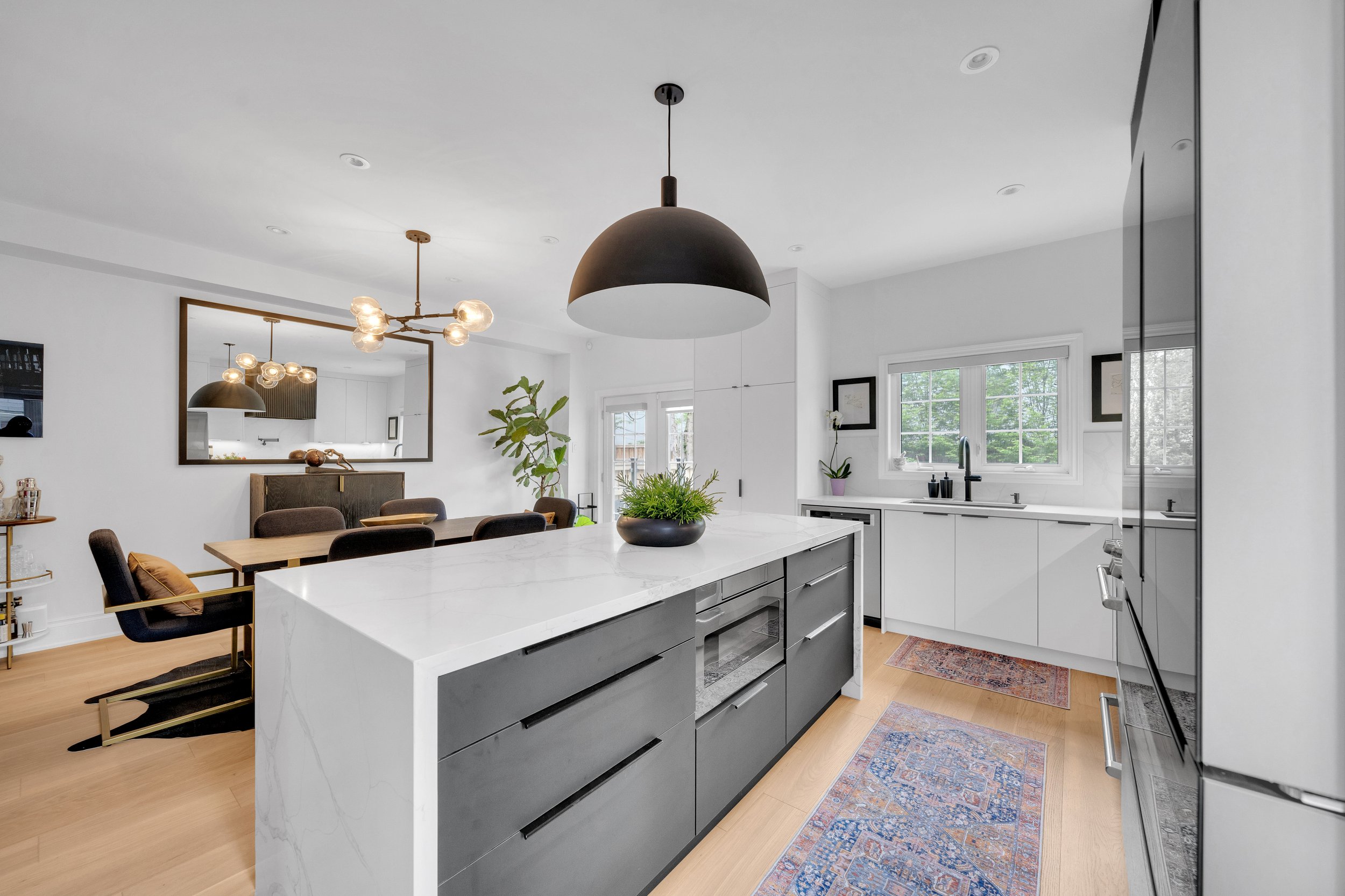 Modern kitchen and dining area with island, black pendant light, dining table, chairs, large mirror, and plants.