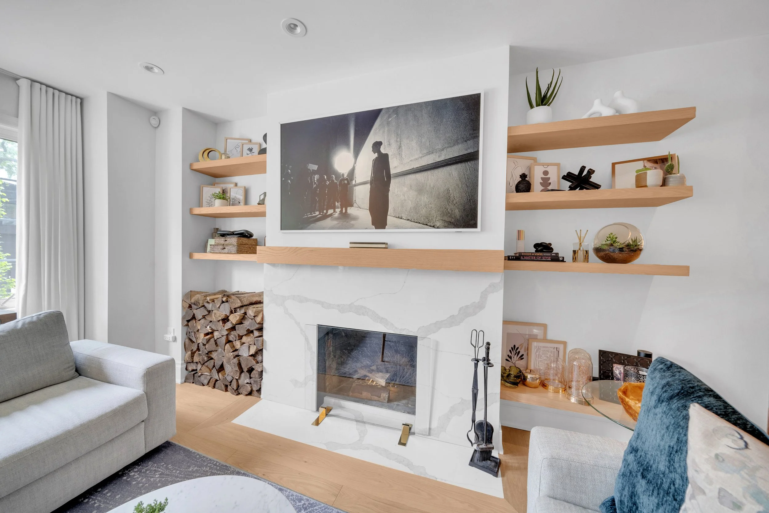 Modern living room with a marble fireplace, wooden mantel, and decorative shelves. Stacked firewood beside the fireplace. Gray sofa and plush blue pillows. Wall-mounted TV displaying a black-and-white photo.