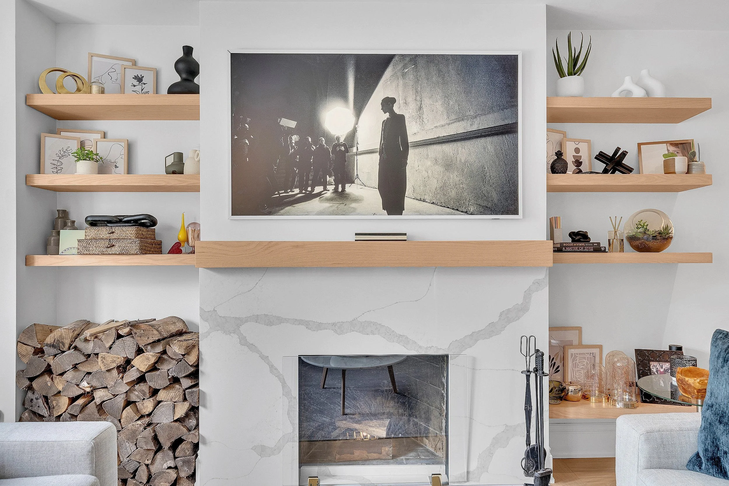 Modern living room with TV above fireplace, wooden shelves with decor, and stacked firewood.