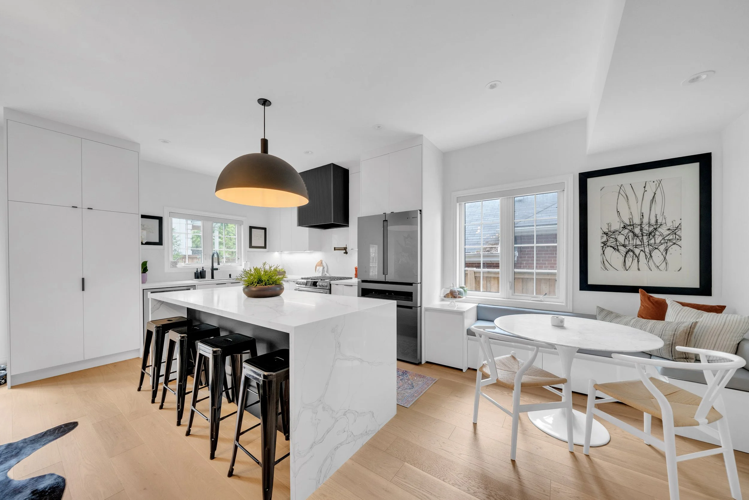 Modern kitchen with white cabinets and island, black metal stools, pendant light, stainless steel appliances, and a round dining table with chairs near a large window.