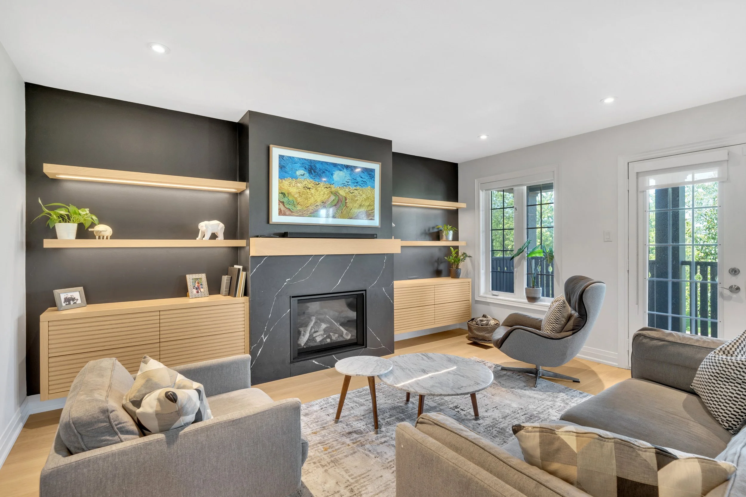 Modern living room with grey sofas, round marble coffee table, black accent wall with fireplace, wooden shelves, and large painting; windows with outdoor view.