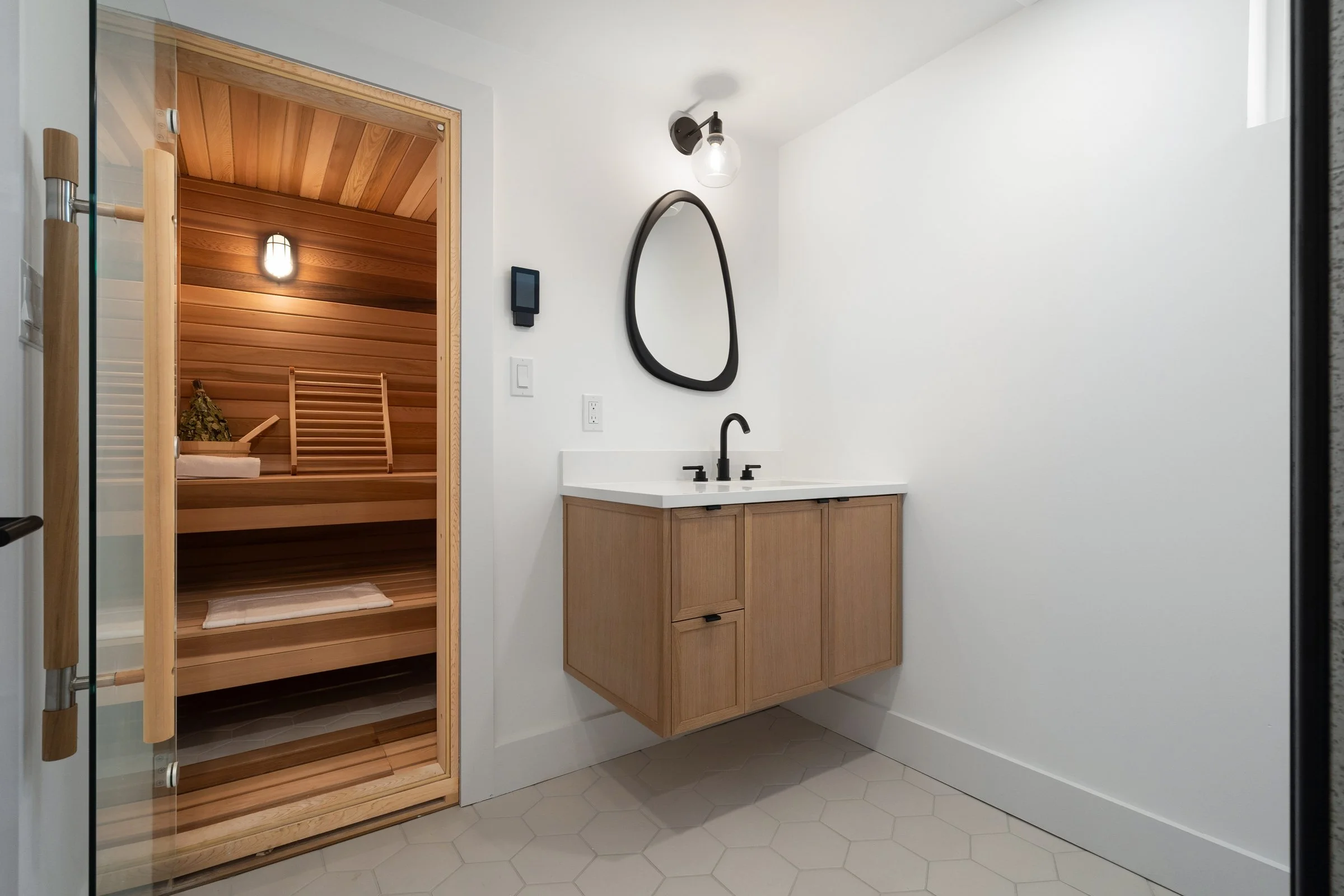 Modern bathroom with a sauna, wooden cabinets, hexagonal tile flooring, and an oval mirror above a sink.