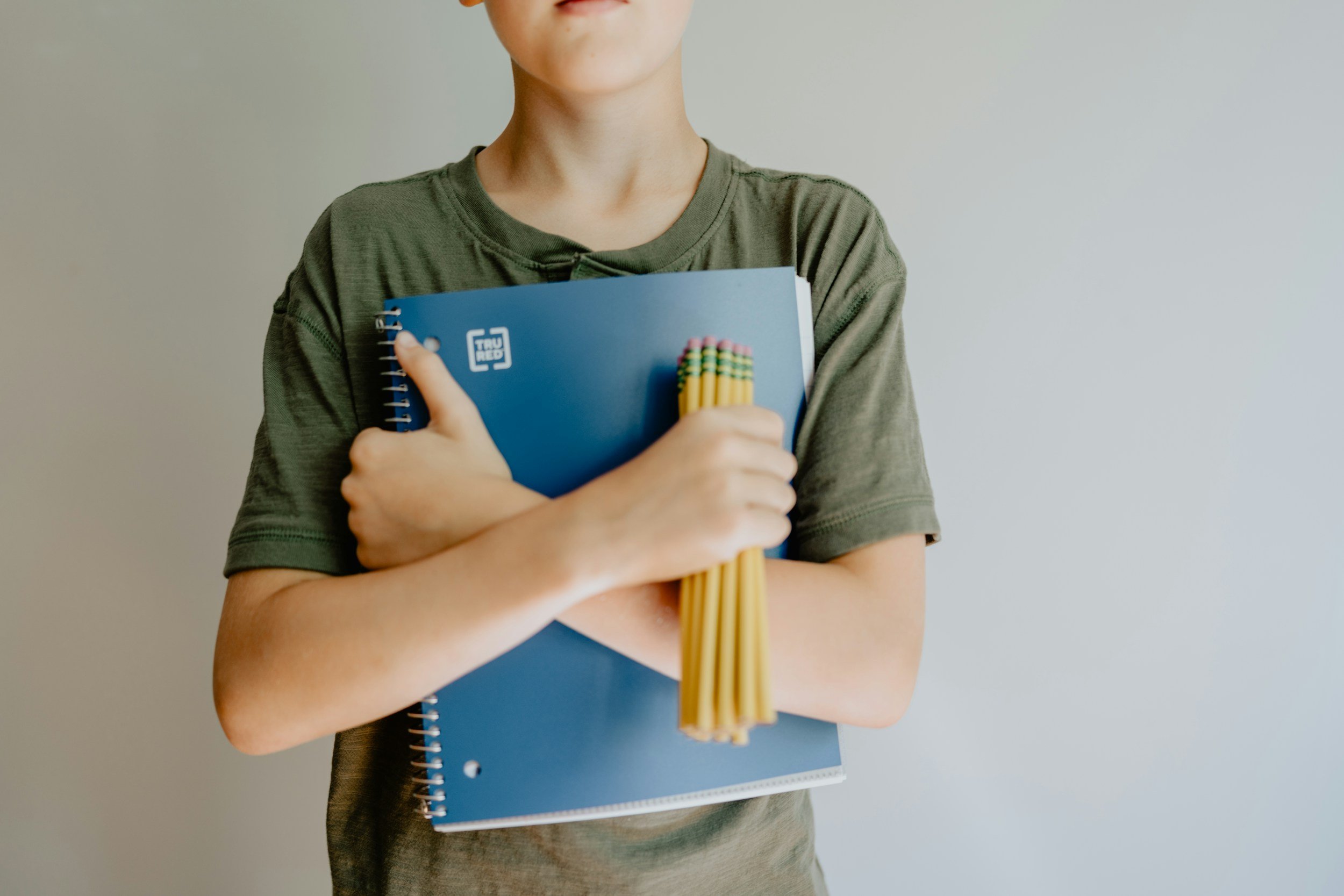 A young man holding a notebook in one hand and a handful of pencils in the other. His arms are crossed in front of his chest. He might be feeling the stress of building out his college list. If so, he could use the help of a True Honors consultant.