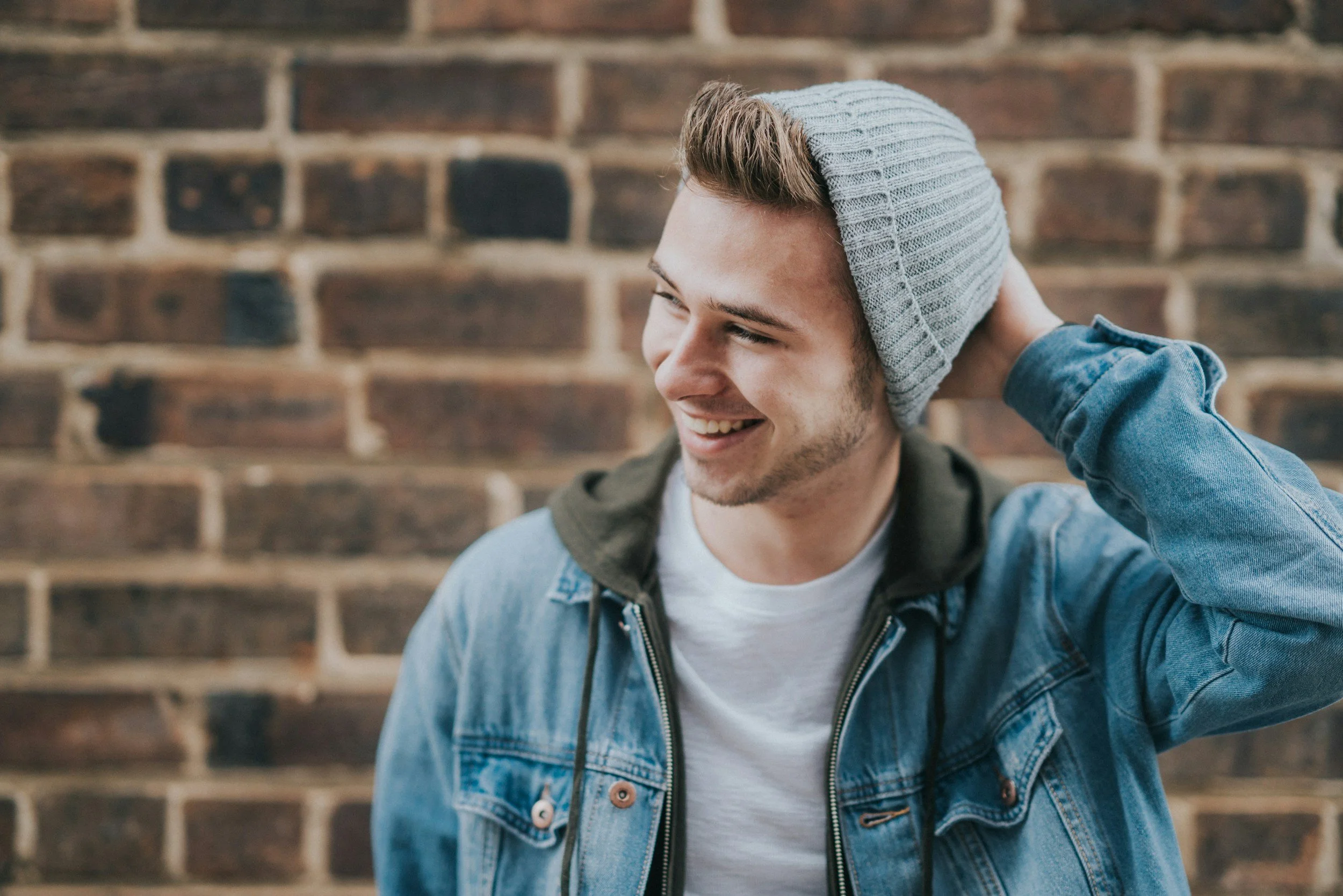 A young man with a beard and short hair smiling, wearing a gray beanie and a blue denim jacket, standing against a brick wall.