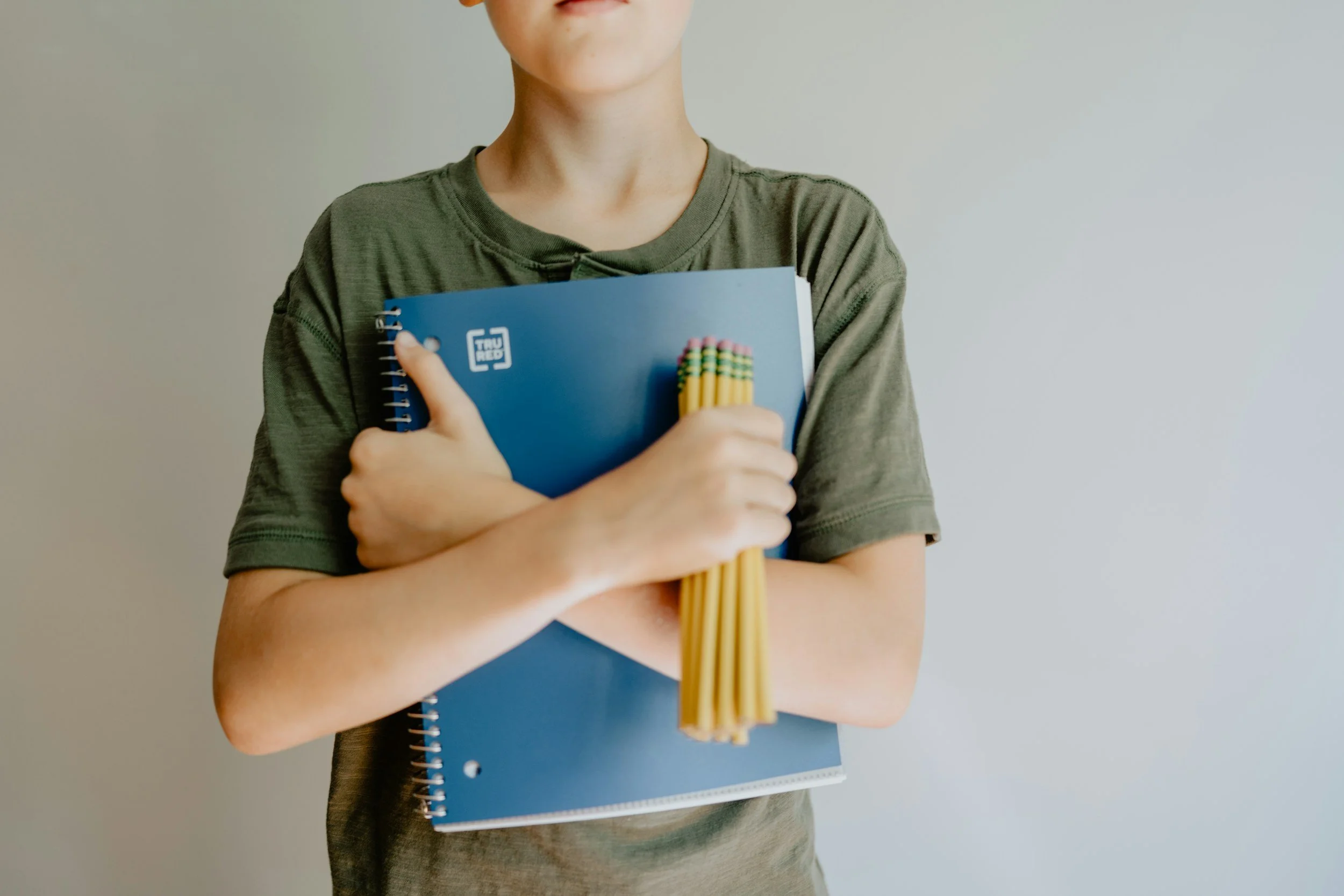 A young man clutching a notebook in one hand and a handful of pencils in the other with his arms crossed across his chest. He appears ready to sit down and find the right school for him.