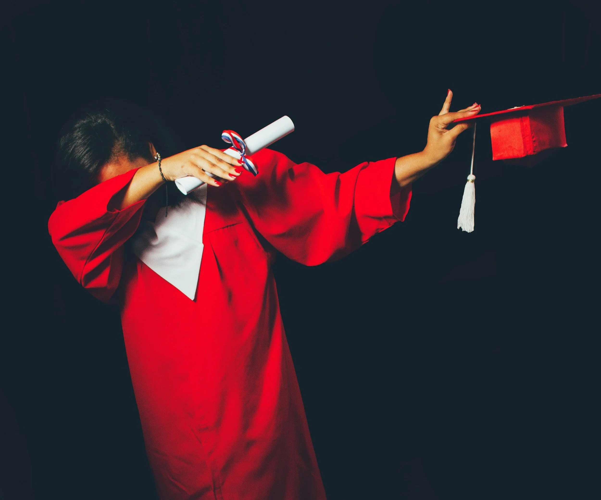 A young person dabbing on stage in a red graduation gown -- their diploma covers their face. This could be you if you work with our expert college counselors for college application assistance.