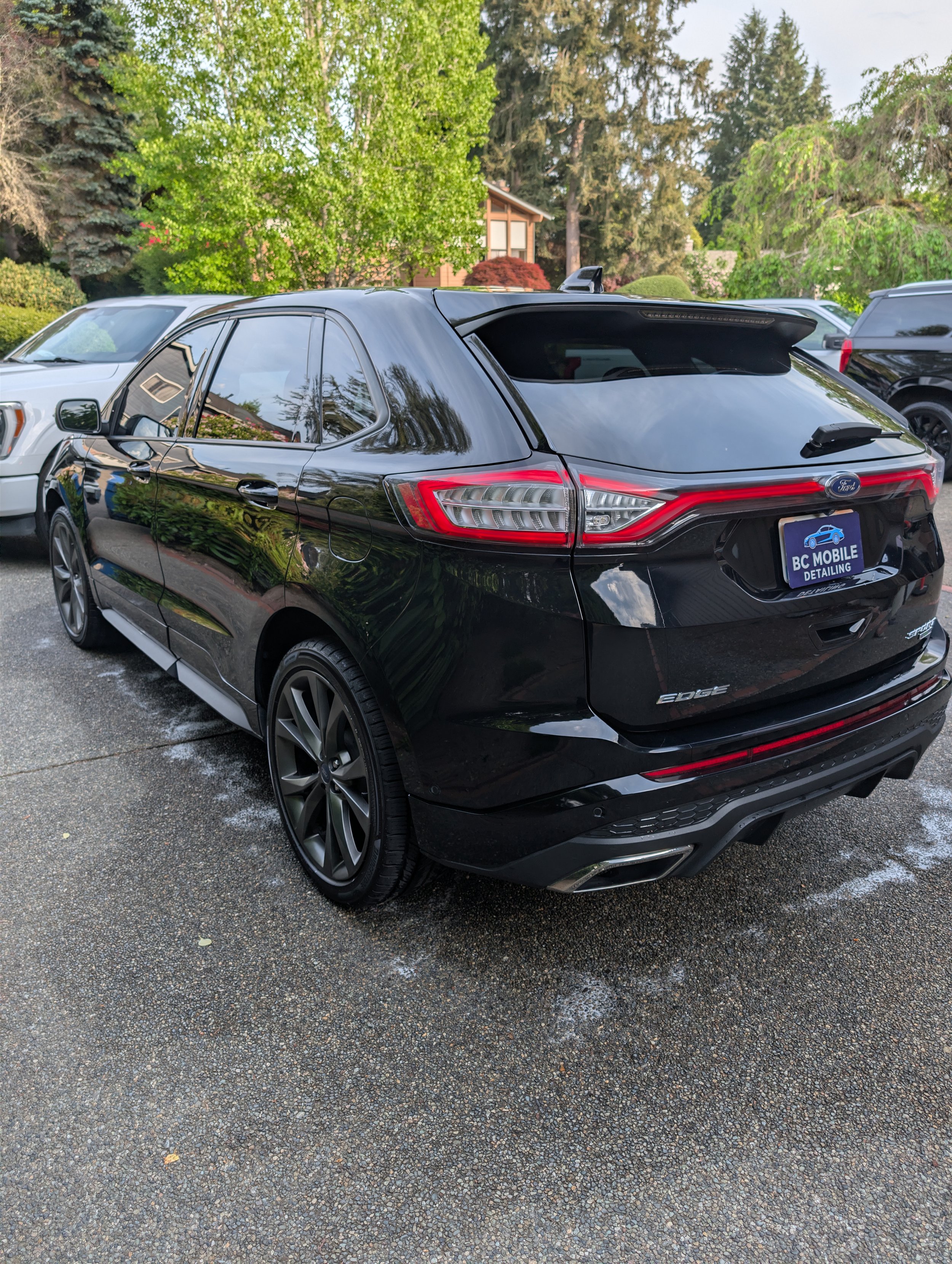 Black Ford Edge SUV parked on asphalt ground with other vehicles visible, surrounded by trees and a small residential house in the background.
