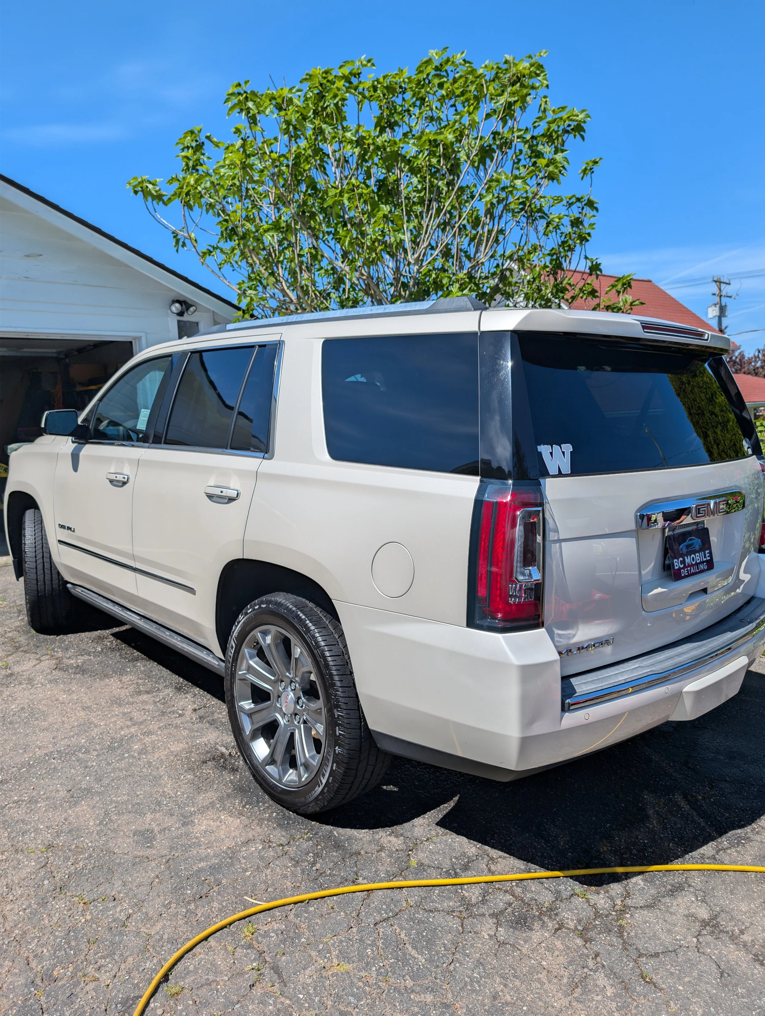 A white GMC SUV parked in front of a house with a tree behind it. The car has a black tinted rear window with a Washington Huskies sticker, and a banner on the back with the text 'BC MOBILE DETAILING.' There is a yellow hose on the asphalt ground in front of the vehicle.