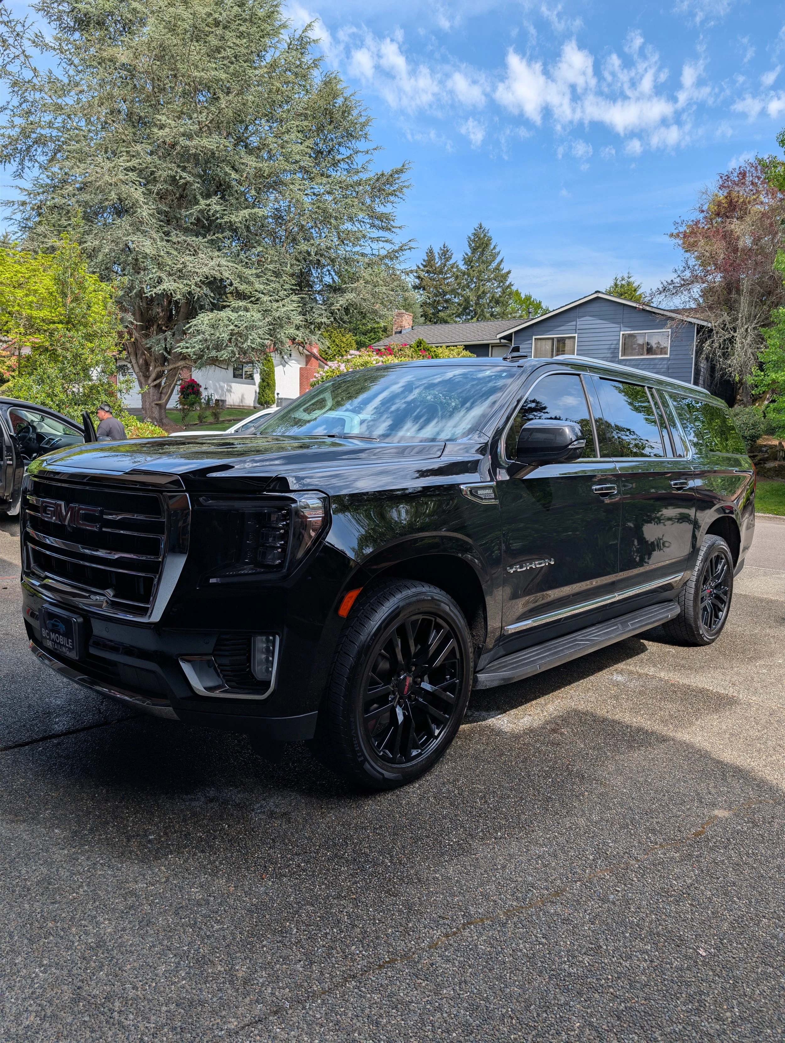Freshly Detailed black GMC Yukon SUV parked on a paved driveway with houses, trees, and a partly cloudy sky in the background.