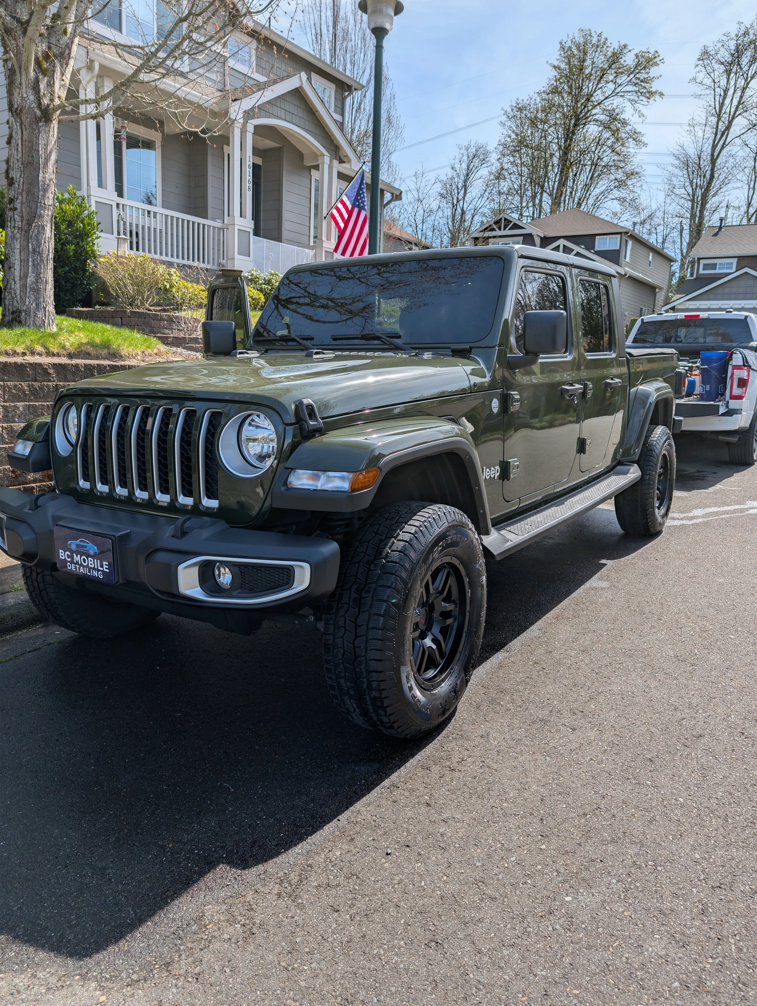 A dark green Jeep parked in front of a residential house with gray siding and white trim. An American flag is displayed near the house, and houses with trees are visible in the background on a sunny day.