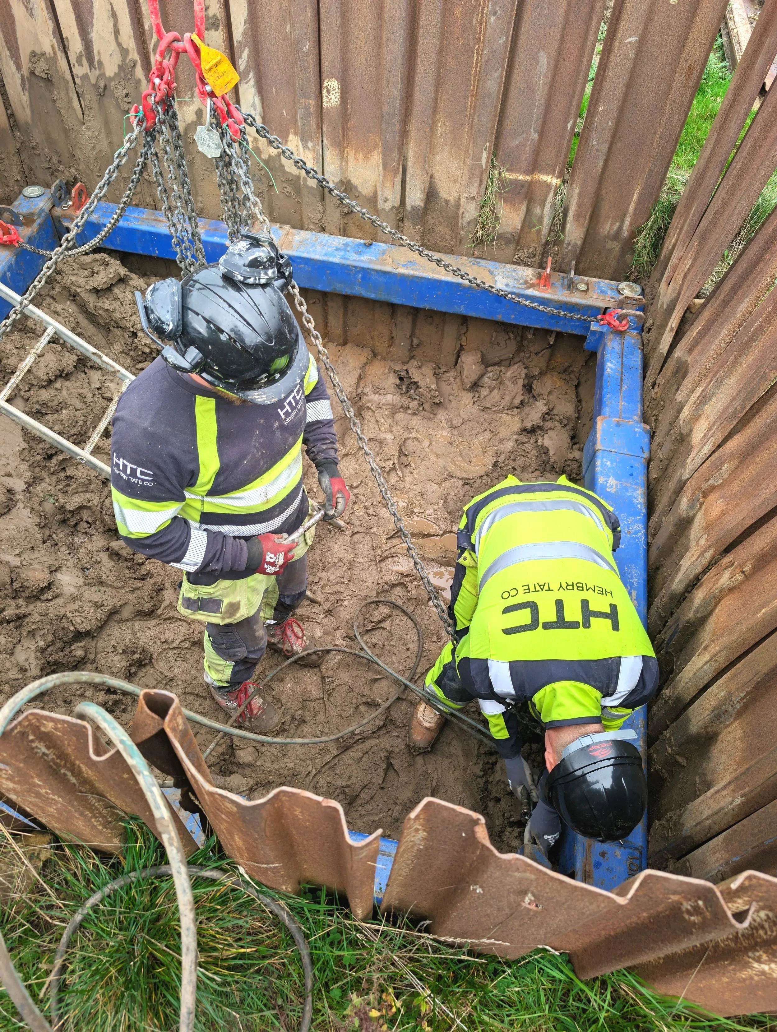 Two construction workers in safety gear working inside a deep excavation site with steel walls.