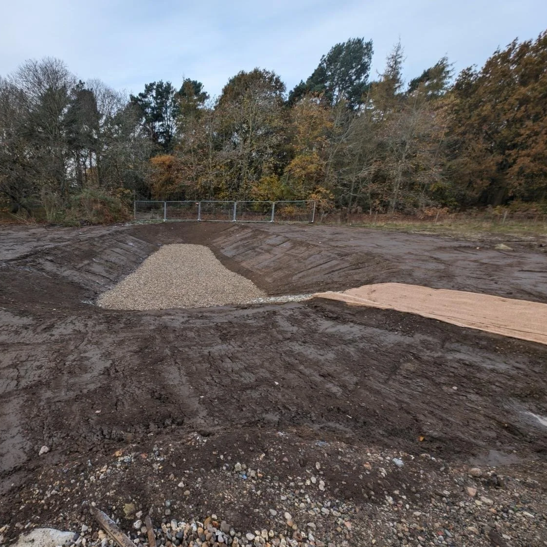 A construction site with a gravel pile and dirt paving, surrounded by trees and a metal fence at the background.