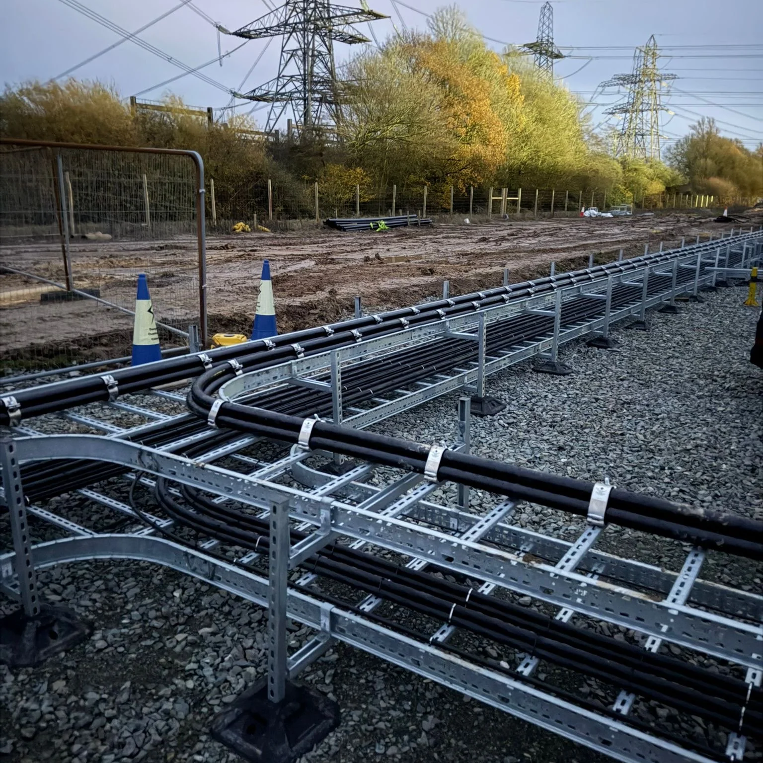Construction site with electrical cable trays, black cables, blue and white safety cones, and power lines in the background, under a partly cloudy sky.