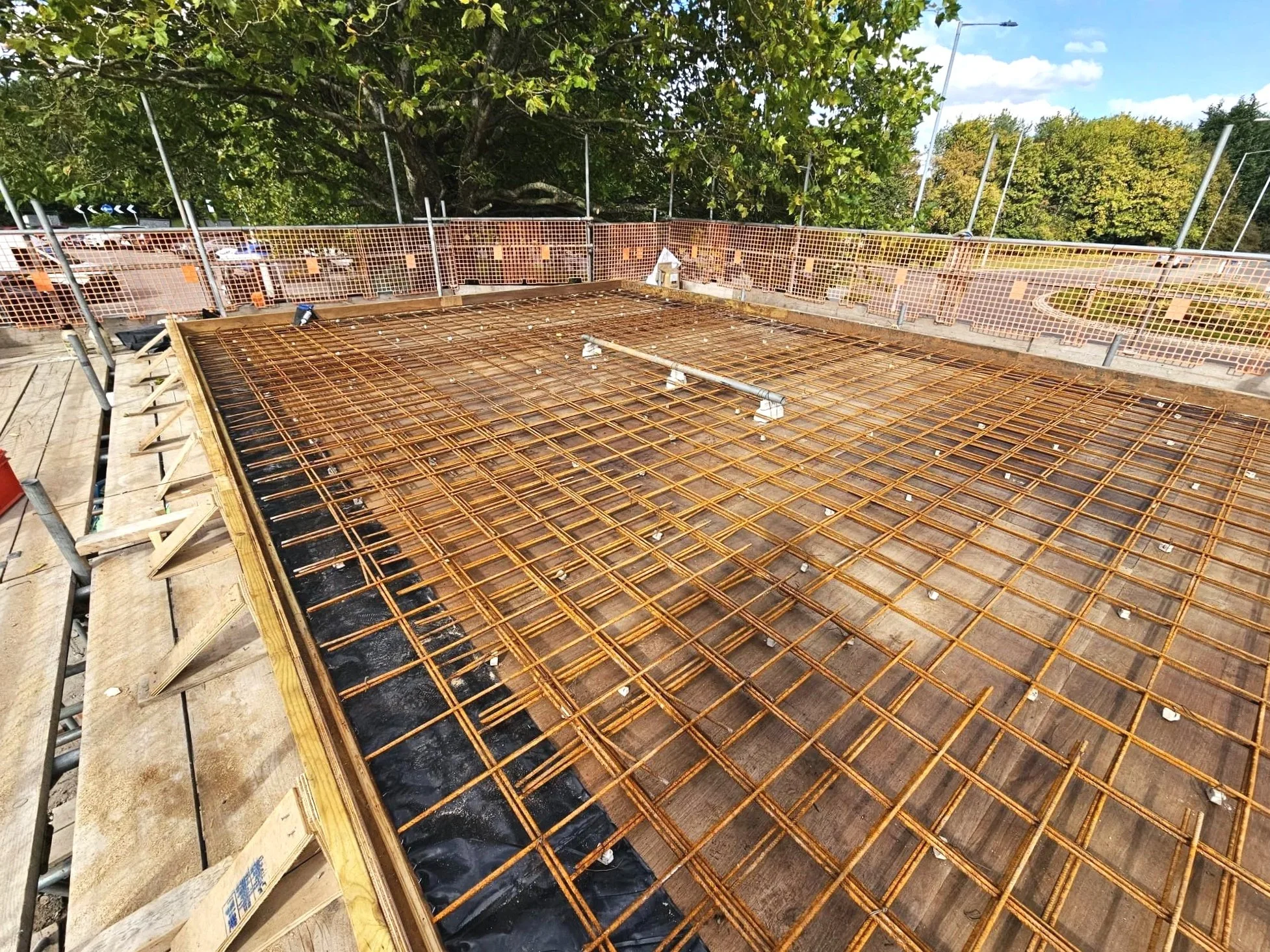 Construction site with steel rebar grid for a concrete slab, surrounded by safety fencing, under a clear blue sky with trees in the background.