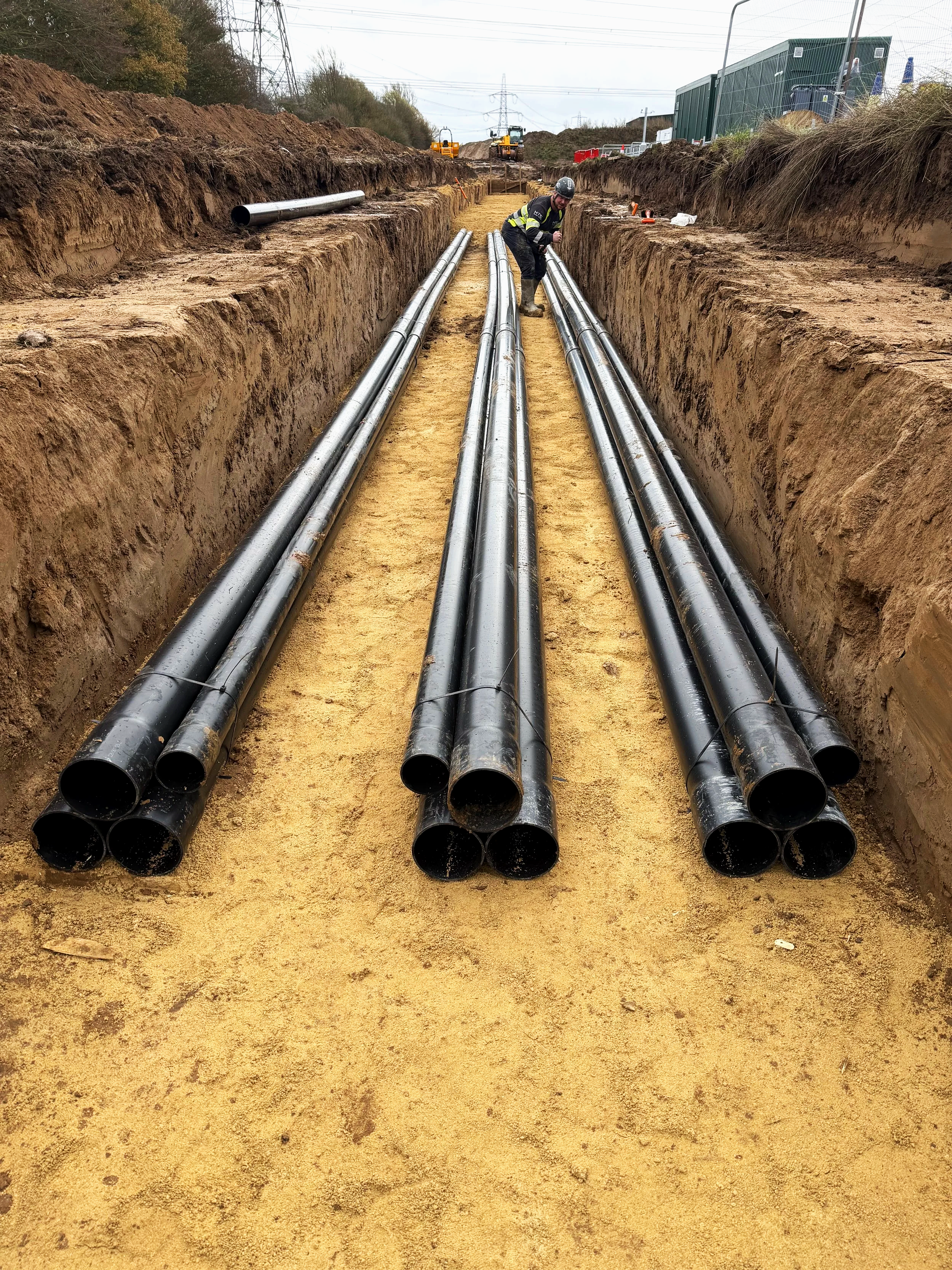 Construction worker inspecting black pipes in a trench on a construction site.
