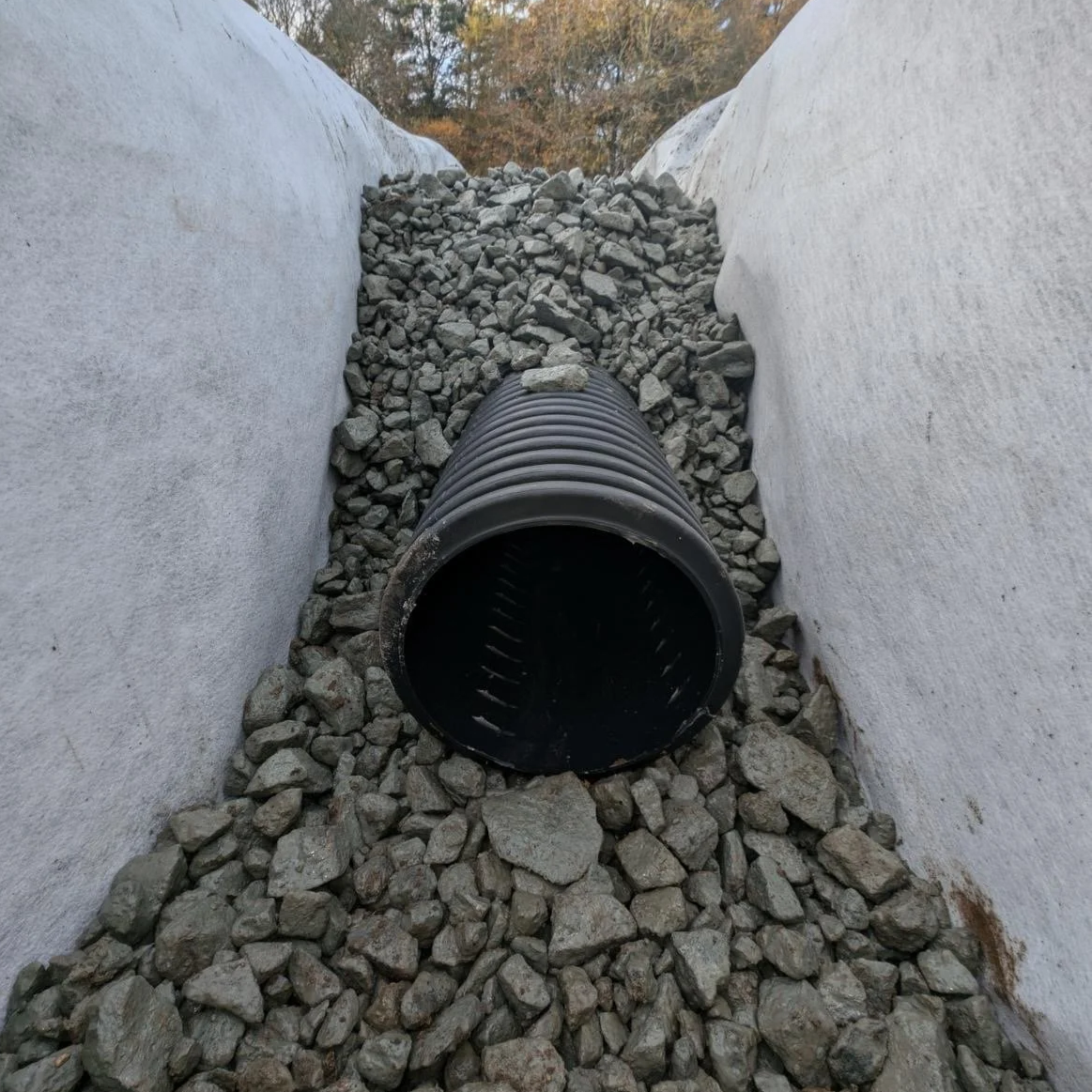 Black corrugated plastic pipe lying in a trench with white concrete walls, filled with rocks and gravel, outdoors with trees in the background.
