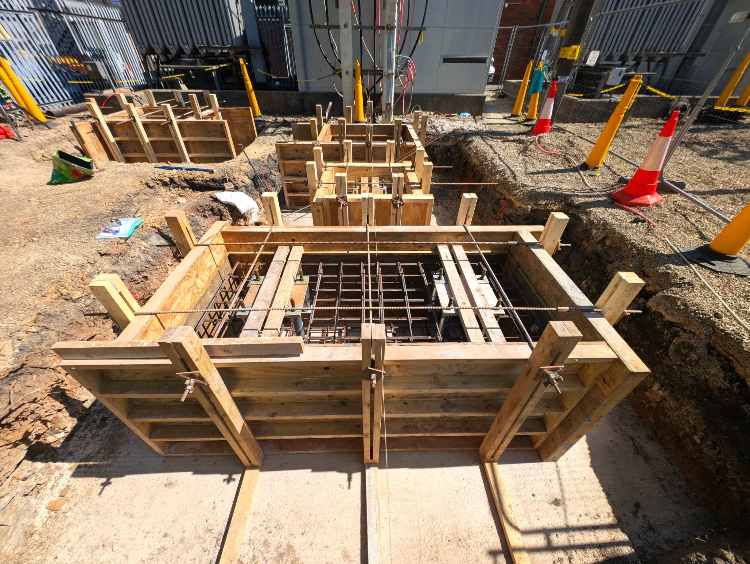Construction site with wooden forms and rebar for concrete foundation, orange safety cones, and fencing around the area.