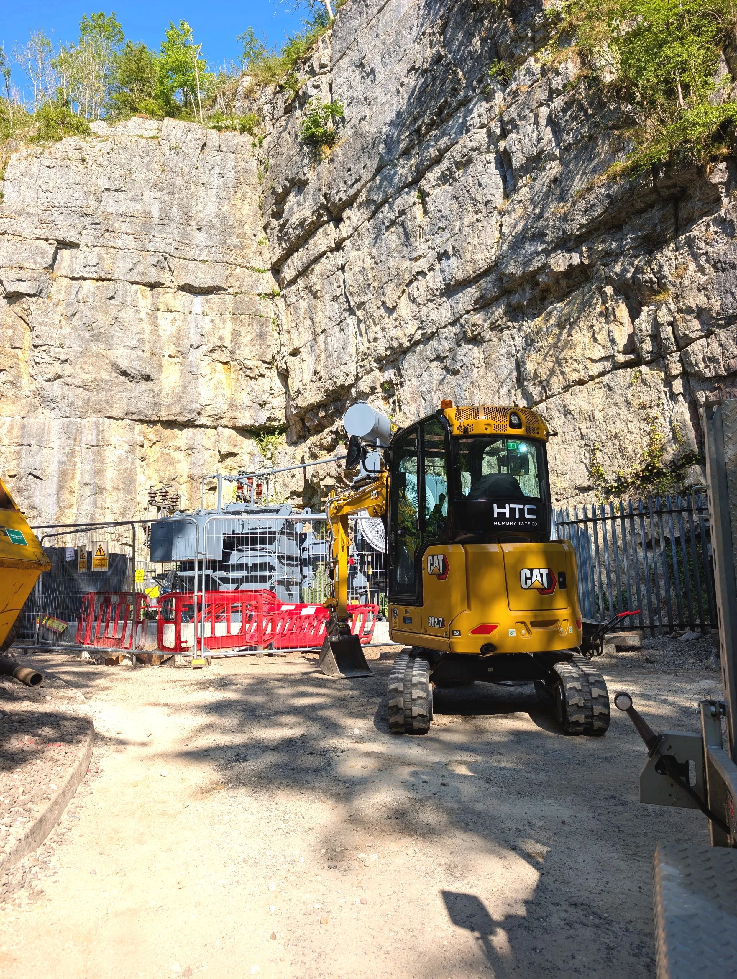 Construction site with a small yellow excavator in front of a rocky cliff, surrounded by safety barriers and equipment.