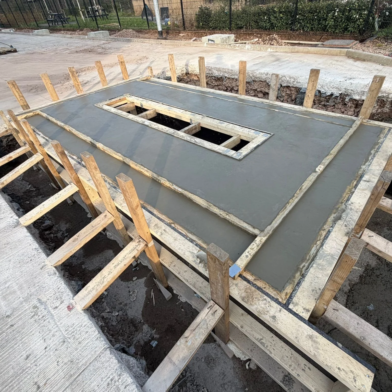 Concrete is being poured into a form to create a foundation for a building, with wooden stakes supporting the formwork.