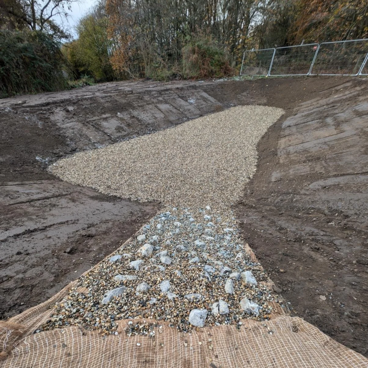 A construction site with earth moving equipment and a gravel bed being prepared for paving, with trees and a temporary fence in the background.