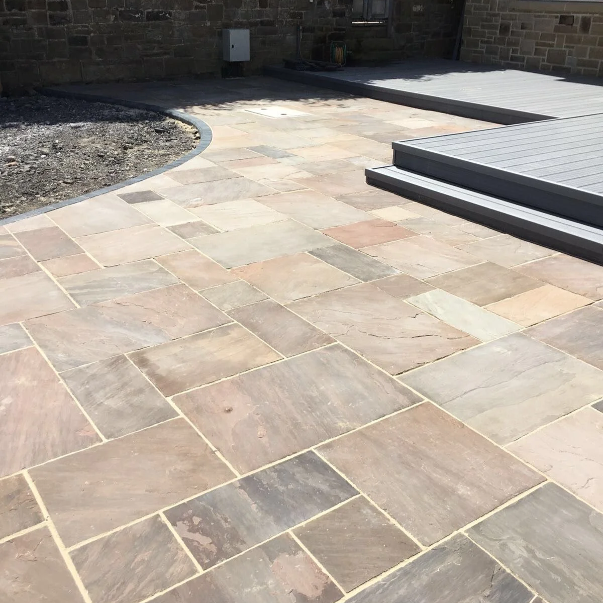 Stone patio with a curved border, adjacent to a gravel area and a raised deck with a ramp.