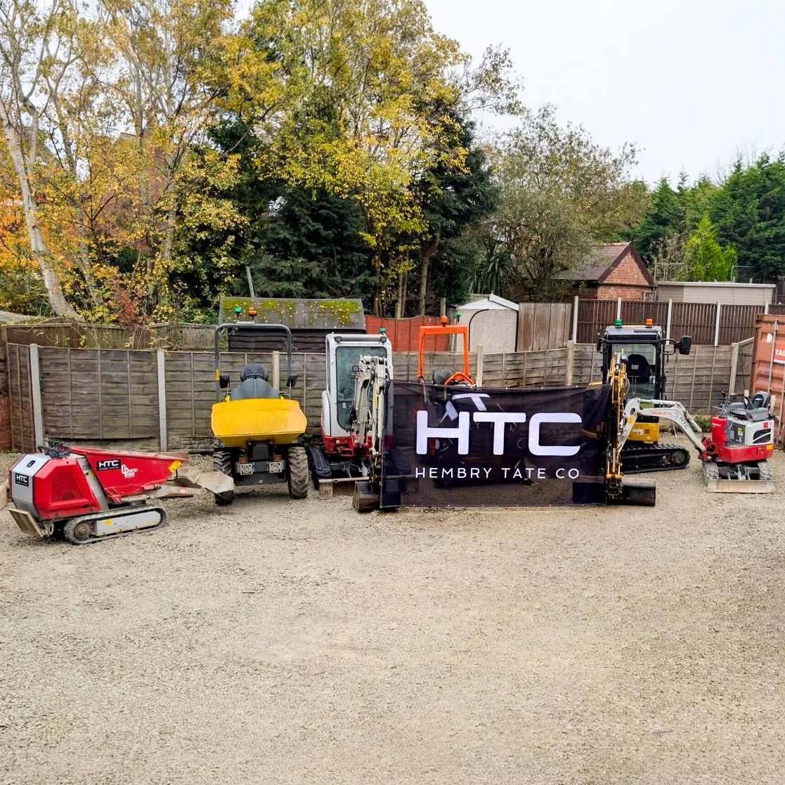 Construction equipment and machinery, including compactors and excavators, with a HTC banner in front, set against a wooden fence and trees with fall foliage.