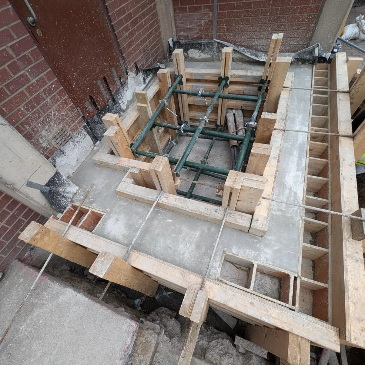 Construction site with wooden formwork and green steel pipes for concrete pouring, surrounding the foundation of a building, next to brick walls.