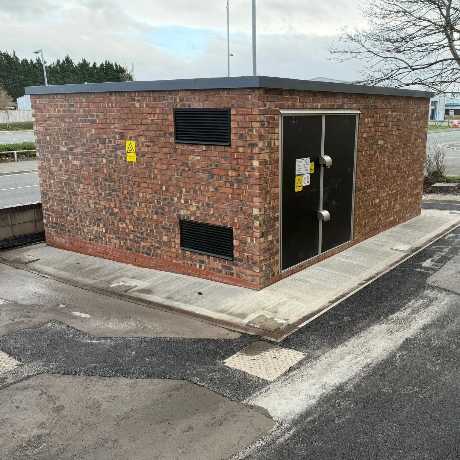 A small brick utility building with black vents and a black door with warning signs, located on a paved sidewalk in a parking lot with street view and cloudy sky.