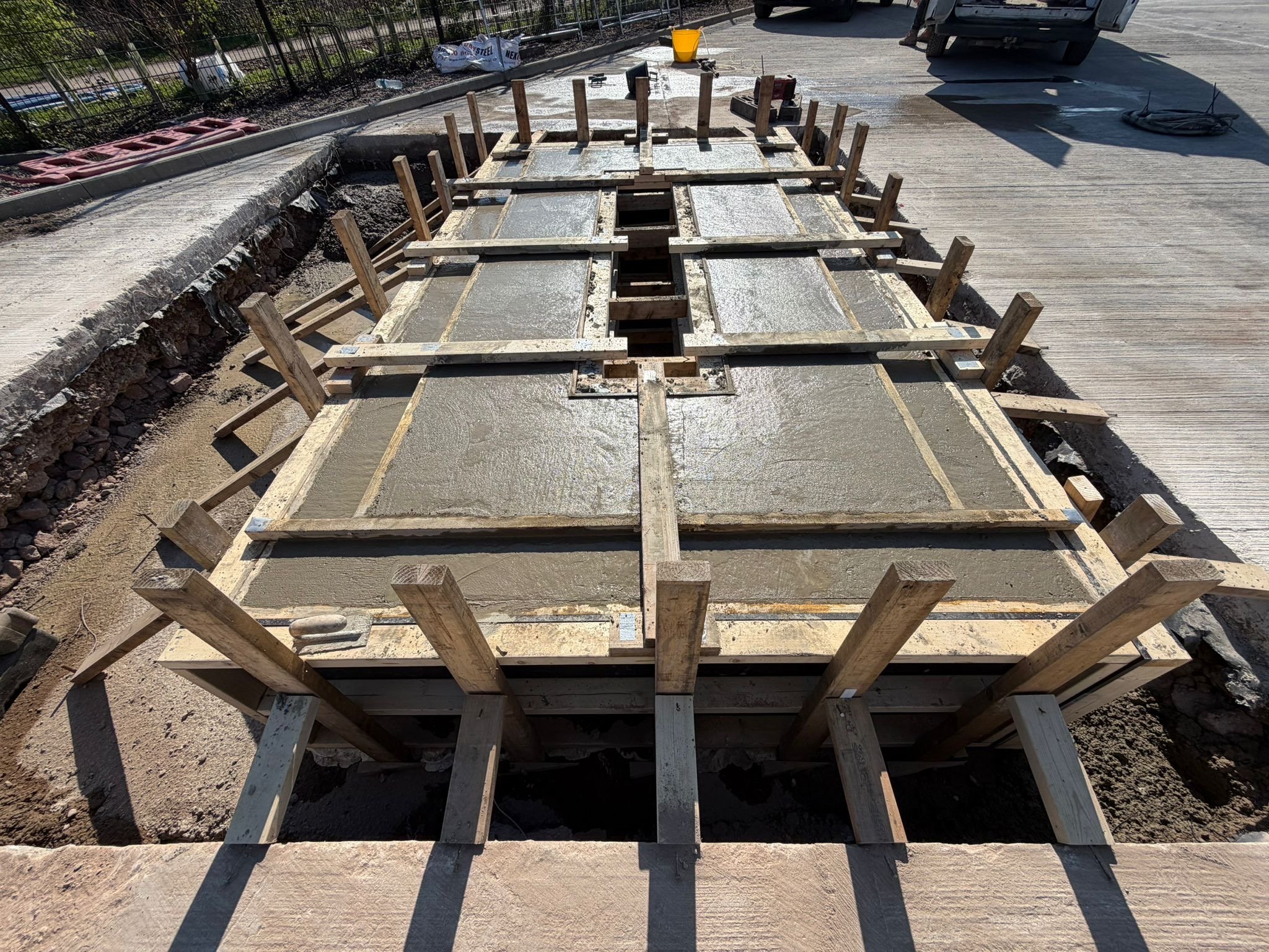 Concrete being poured into a form for a sidewalk or pavement, with wooden and metal framing for support, on a construction site.