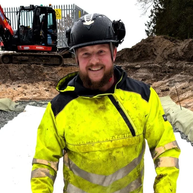 Construction worker wearing a yellow safety jacket and a black helmet, smiling at a construction site with equipment in the background.